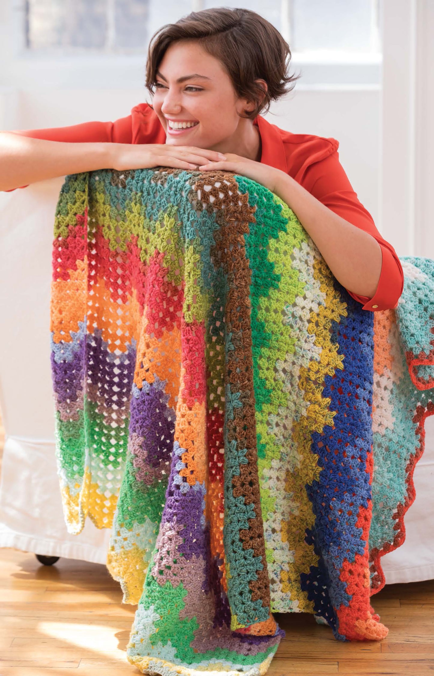 A smiling woman in a red shirt leans on a white couch, showcasing a vibrant zigzag crocheted blanket made with Cakery Crochet by Leisure Arts—an ideal choice if you're seeking beginner crochet patterns.