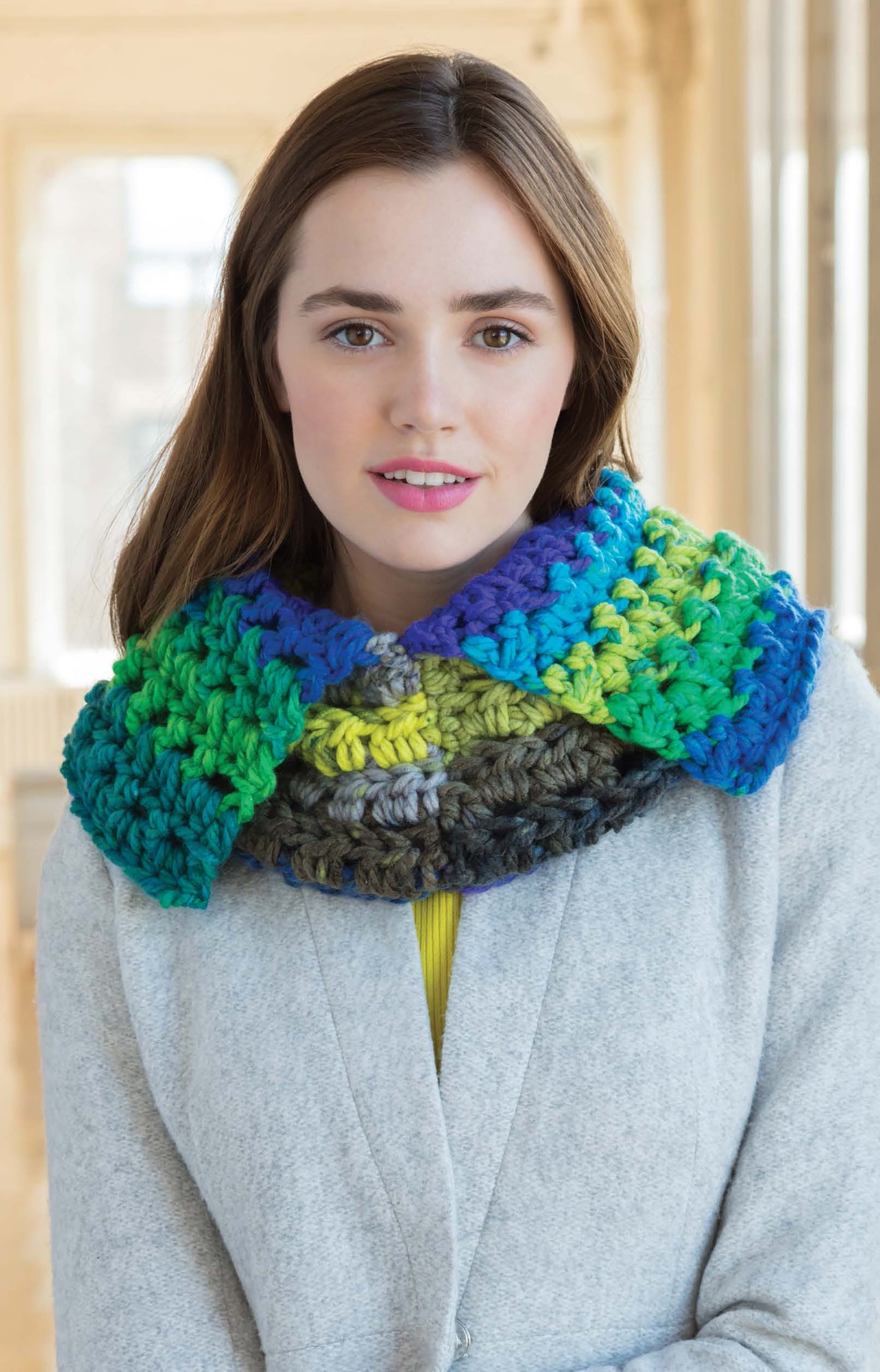 A woman with long brown hair wears a light gray coat and a chunky, multicolored scarf crocheted from Leisure Arts Cakery Crochet yarn in blue, green, and gray tones, standing indoors with natural light behind her.
