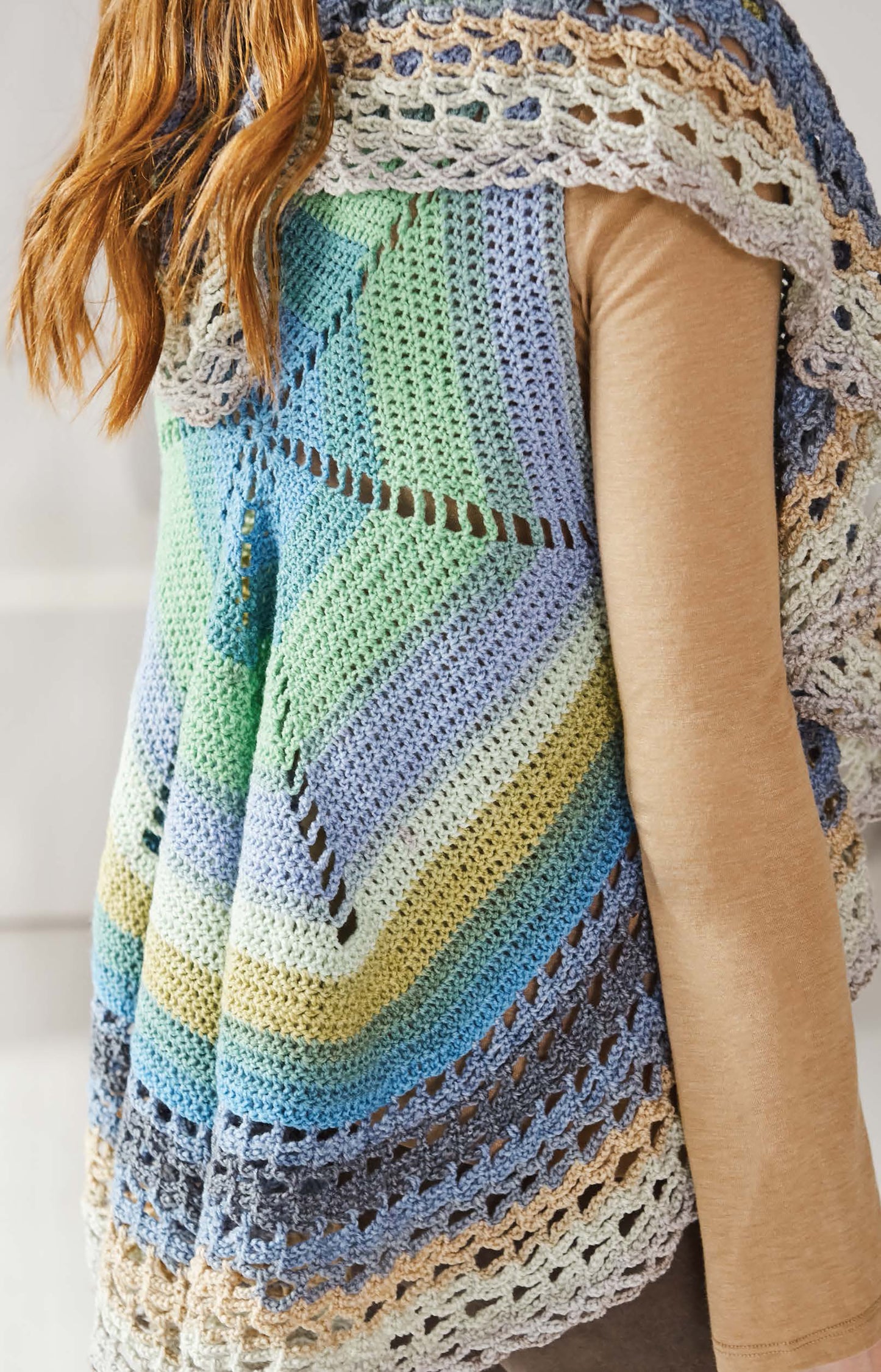 A person with long, light brown hair wears a crocheted vest made from Leisure Arts Cakery Crochet yarn in blue, green, yellow, and cream stripes over a beige long-sleeve shirt, shown from the back and side.
