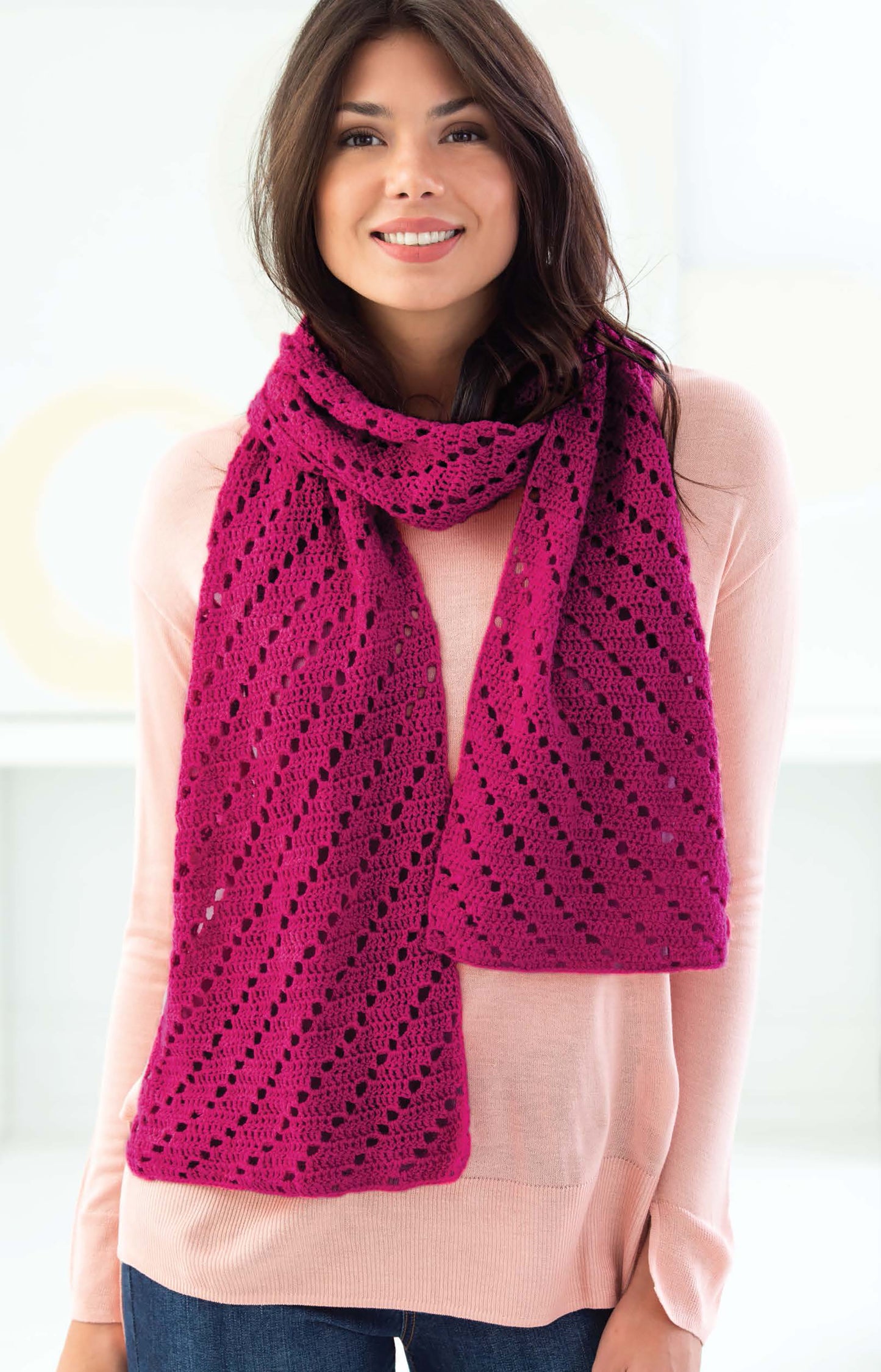 A woman with long brown hair smiles while wearing a pink lace-patterned crochet scarf made with Leisure Arts Cakery Crochet yarn, paired with a light pink sweater and jeans. She stands indoors against a bright, neutral background.
