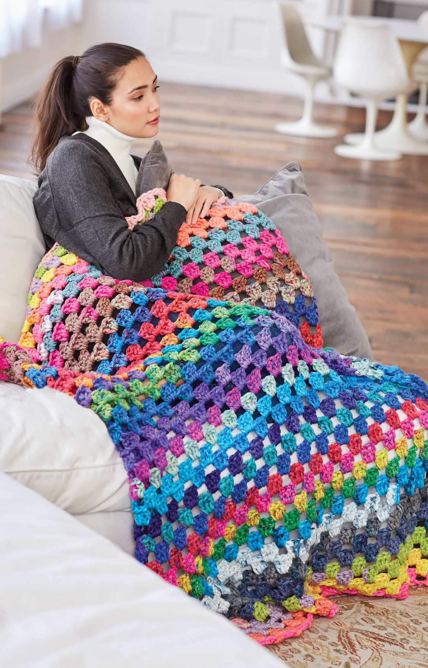 A woman relaxes on a couch, wrapped in a vibrant granny square blanket made with Cakery Crochet by Leisure Arts—perfect for anyone enjoying step-by-step crochet projects in a cozy, modern living room.