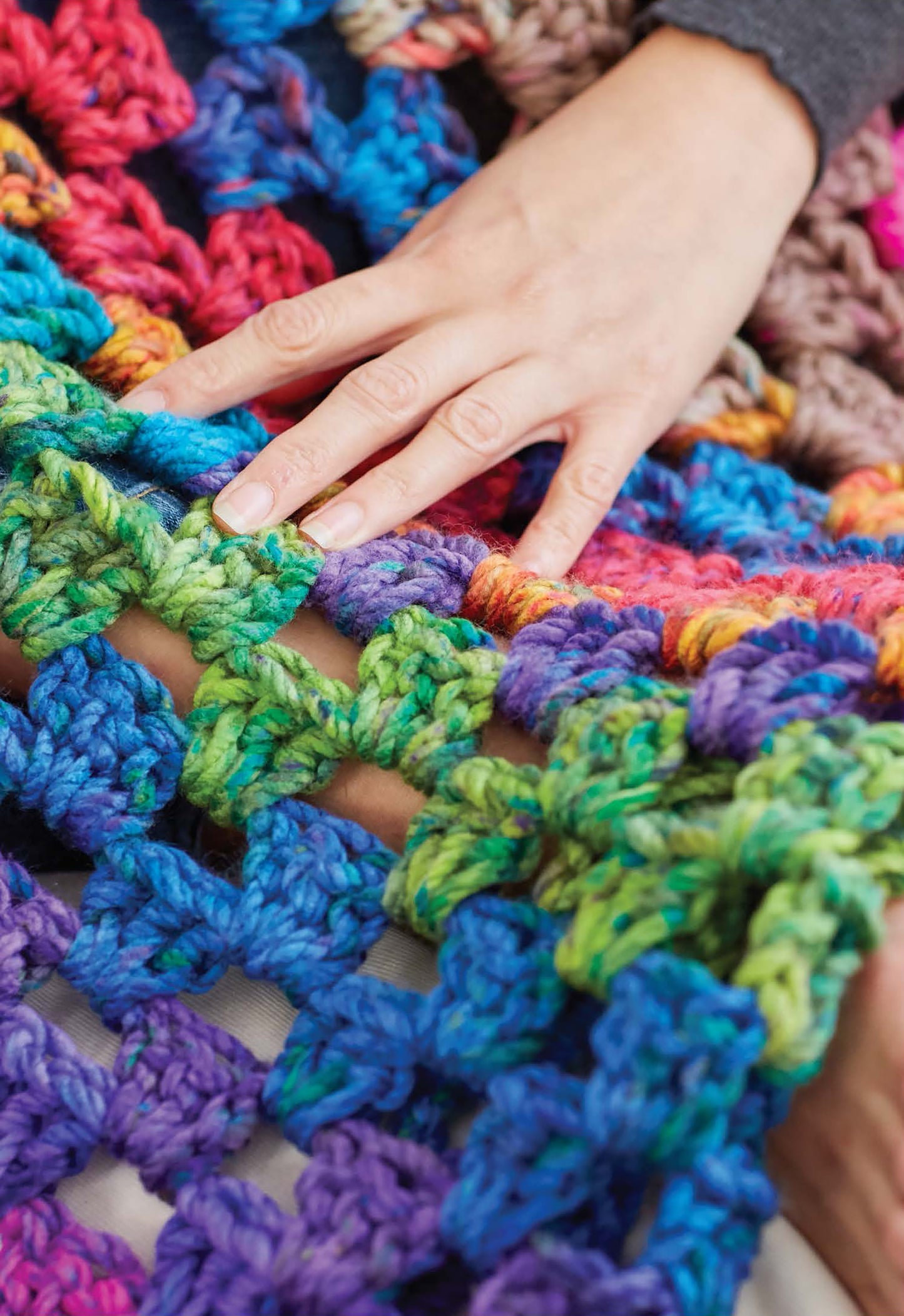 A hand rests on a vibrant, chunky Cakery Crochet blanket by Leisure Arts, featuring thick yarn in blue, green, purple, red, and pink shades with an open lacy stitch—ideal for beginner crochet patterns.