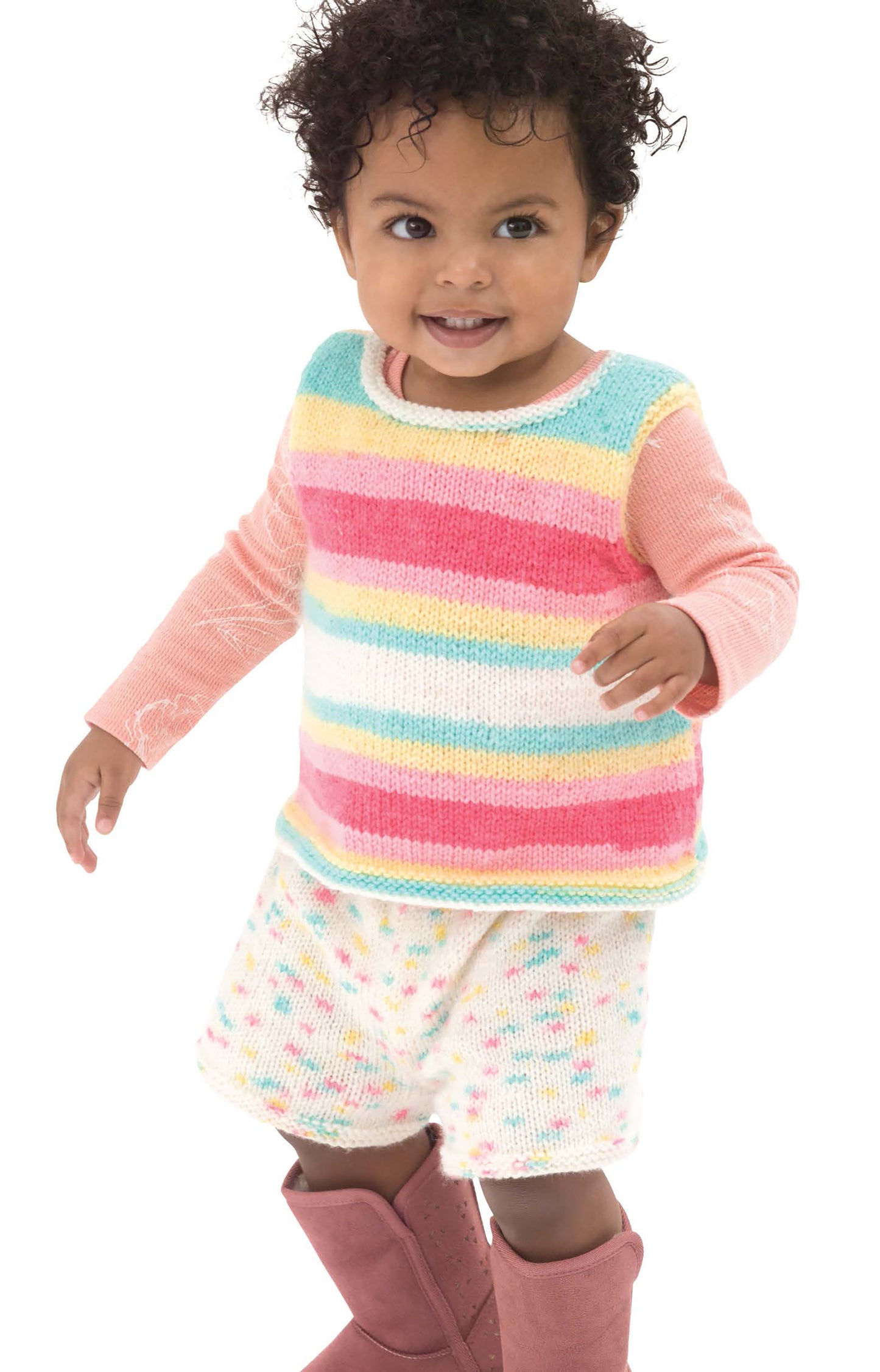 A toddler with curly hair models pastel striped knits and speckled shorts, showcasing the perfect inspiration for handmade baby clothes featured in "Sweet Knits—9 Baby Projects" by Leisure Arts, ideal for beginner knitters.