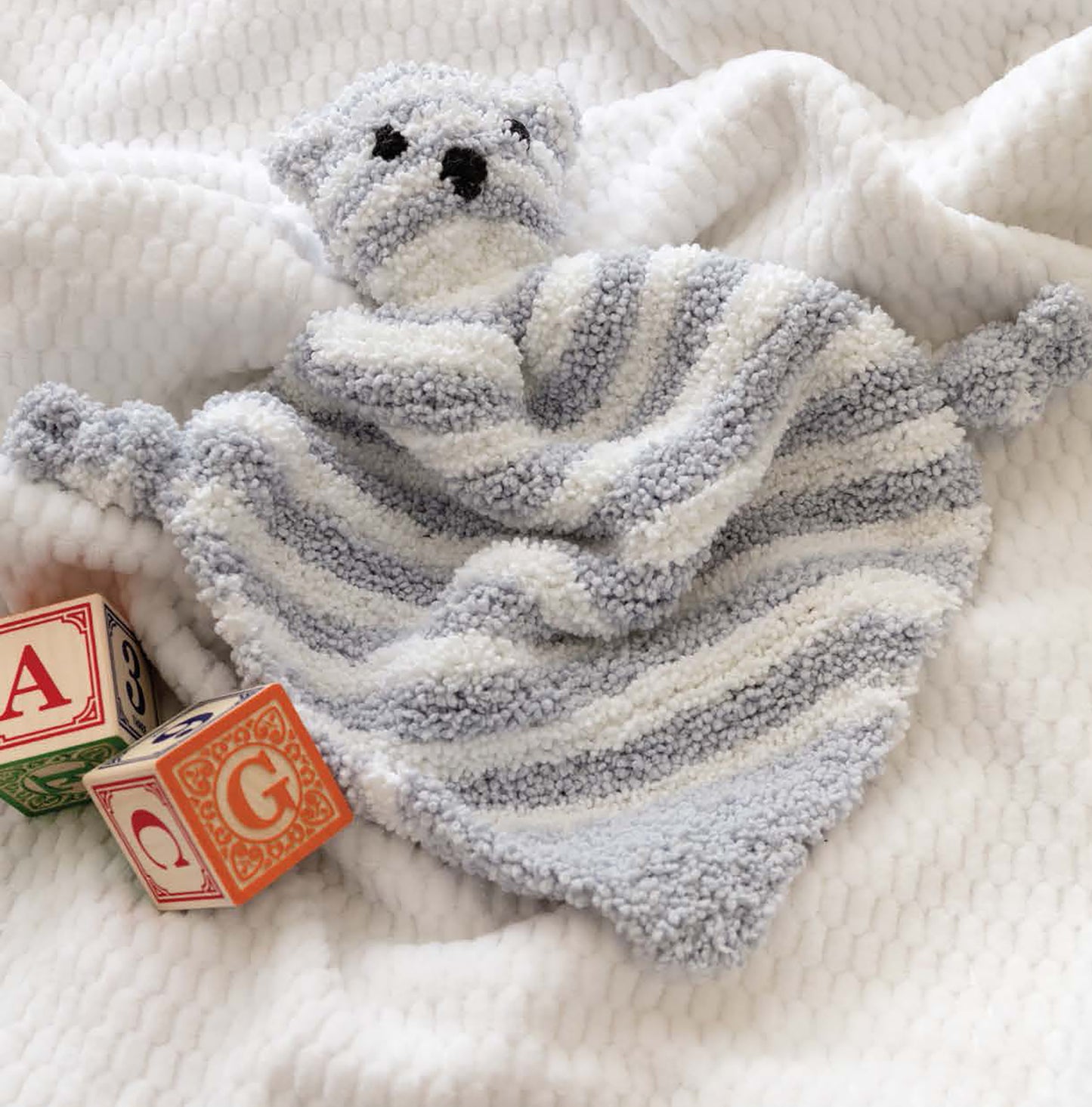The Sweet Knits—9 Baby Projects book by Leisure Arts features a soft, striped gray and white teddy bear lovey blanket, shown on a textured white surface beside wooden alphabet blocks with red Q and orange G.