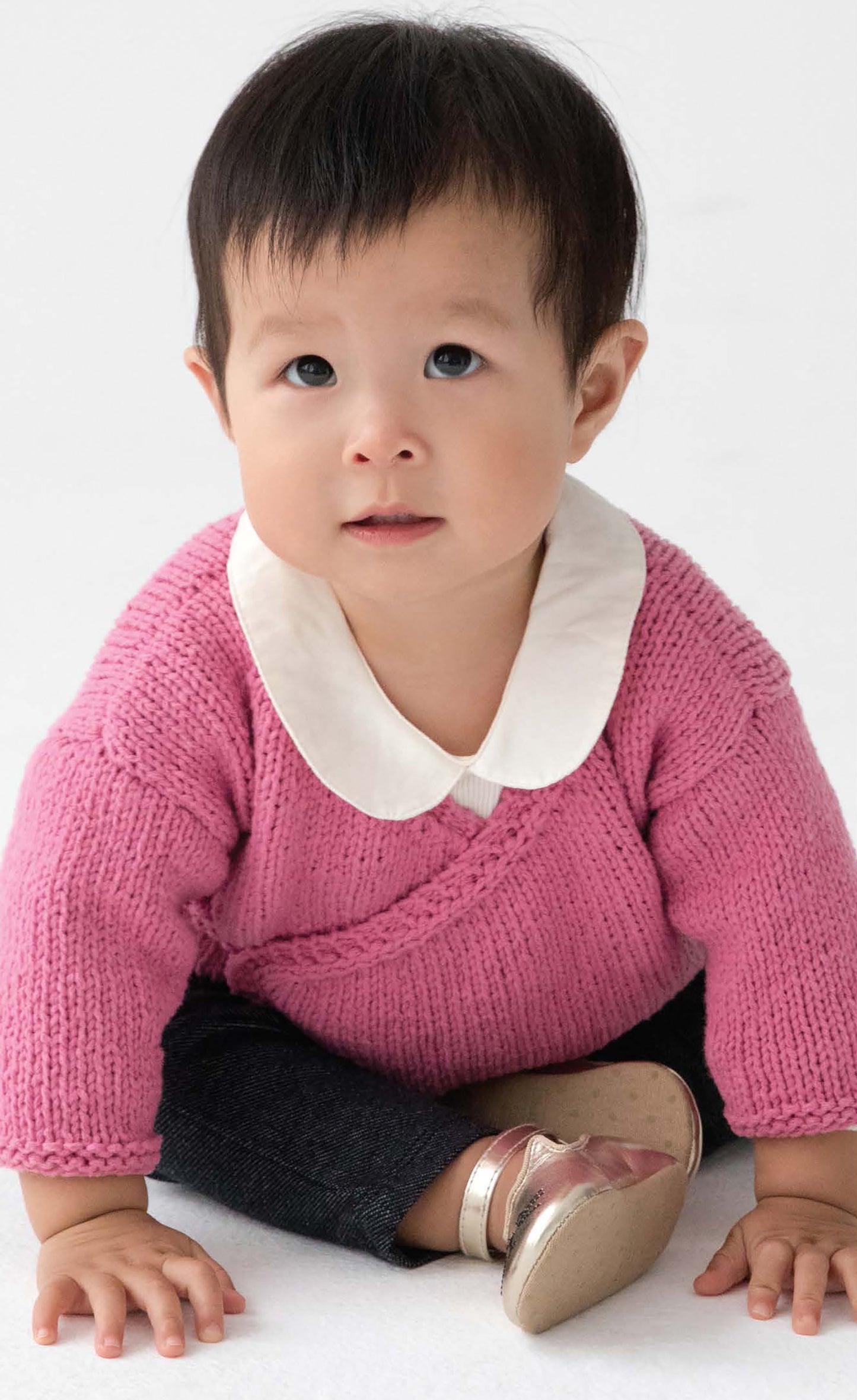 A toddler in a pink knit sweater with a white collar from Leisure Arts’ Sweet Knits—9 Baby Projects sits on the floor in dark pants and shiny shoes, looking up with a neutral expression against a plain background.