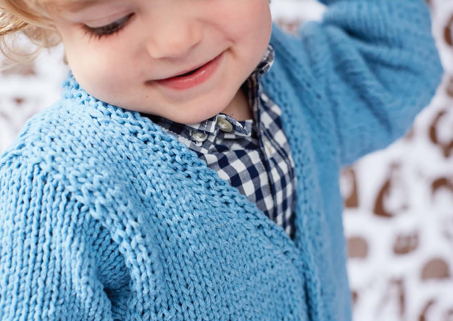 A young child smiles with eyes closed in a close-up, wearing a blue sweater made from Leisure Arts' Sweet Knits—9 Baby Projects, layered over a blue-and-white checkered shirt—ideal for beginner knitters.