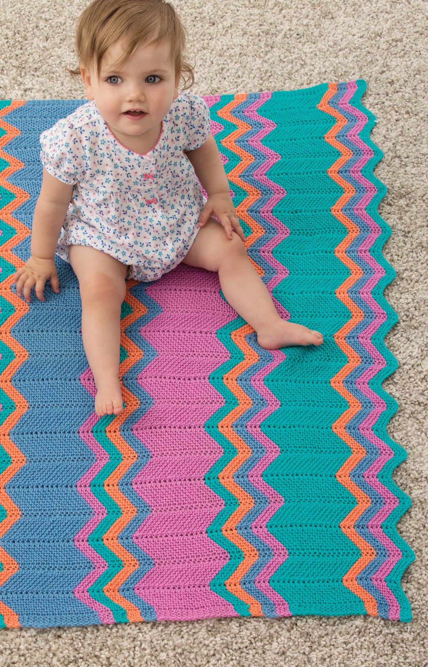 A baby in handmade clothes from Sweet Knits—9 Baby Projects by Leisure Arts sits on a textured carpet and a colorful crocheted zigzag blanket, looking up with an open-mouthed expression.