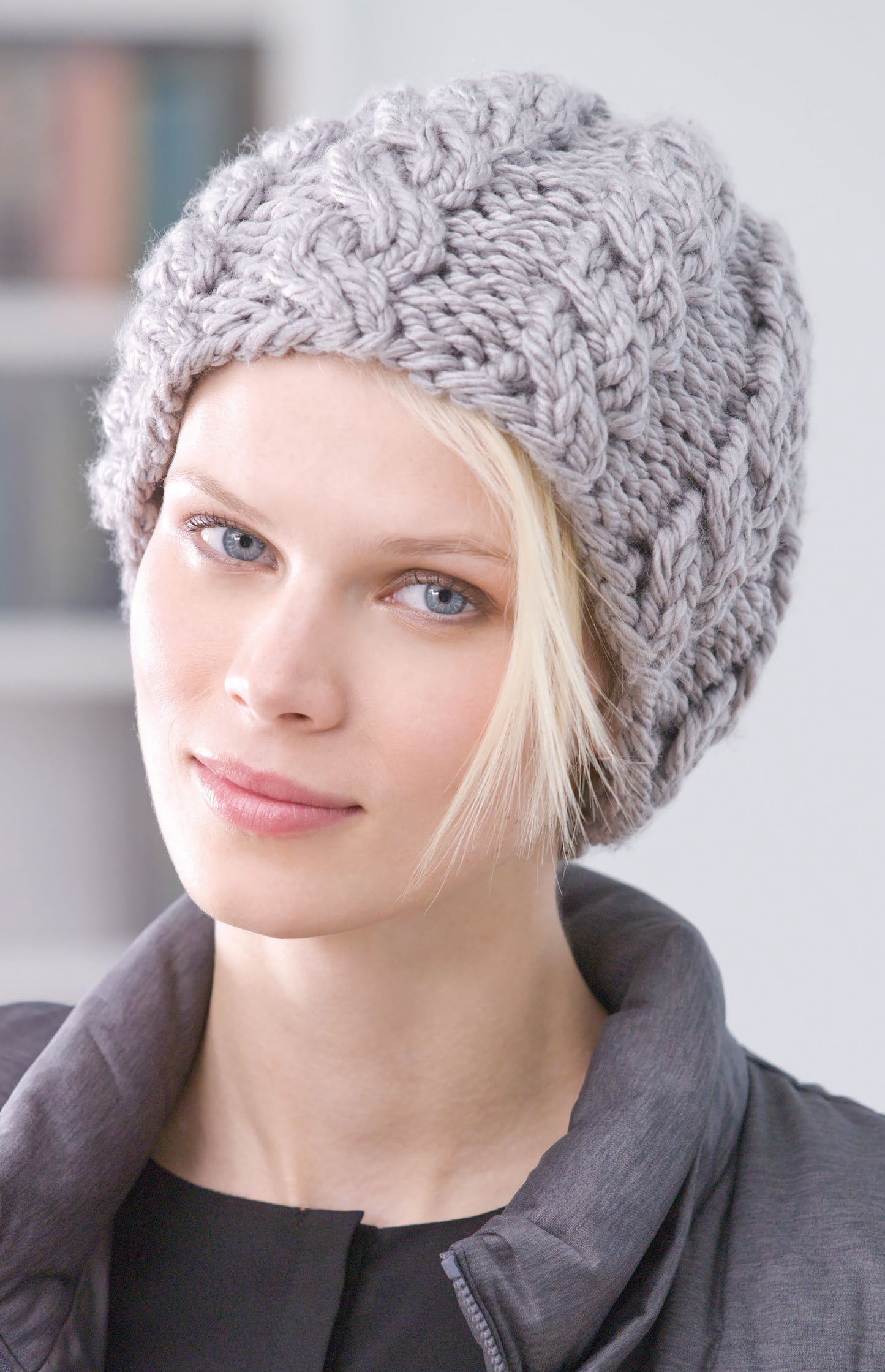 A woman with light skin and blue eyes wears a thick, chunky-knit light gray beanie from Leisure Arts' All Your Favorite Hats collection, paired with a dark jacket and a strand of blonde hair on her forehead.