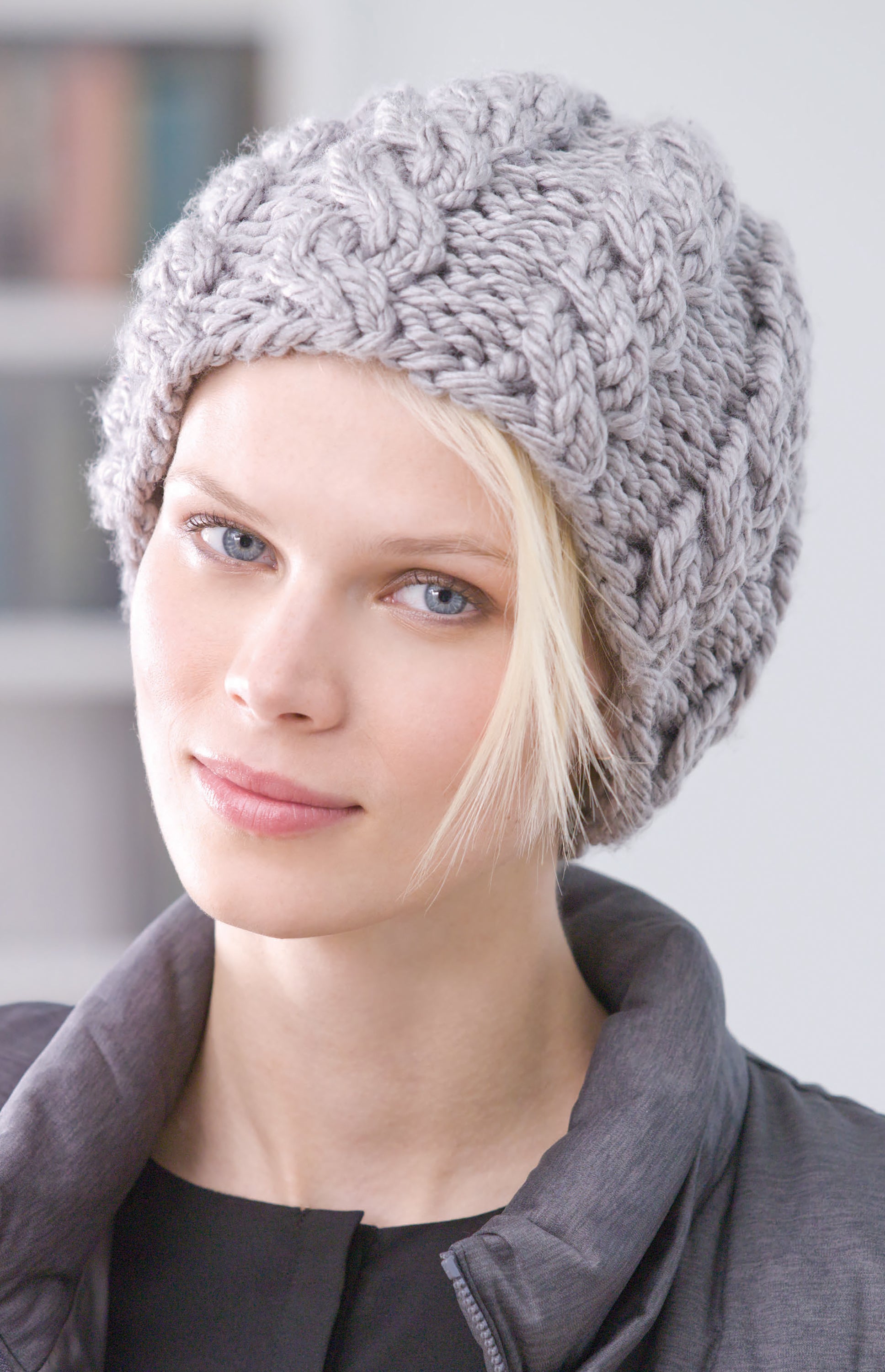 A woman with light skin and blue eyes wears a thick, chunky-knit light gray beanie from Leisure Arts' All Your Favorite Hats collection, paired with a dark jacket and a strand of blonde hair on her forehead.