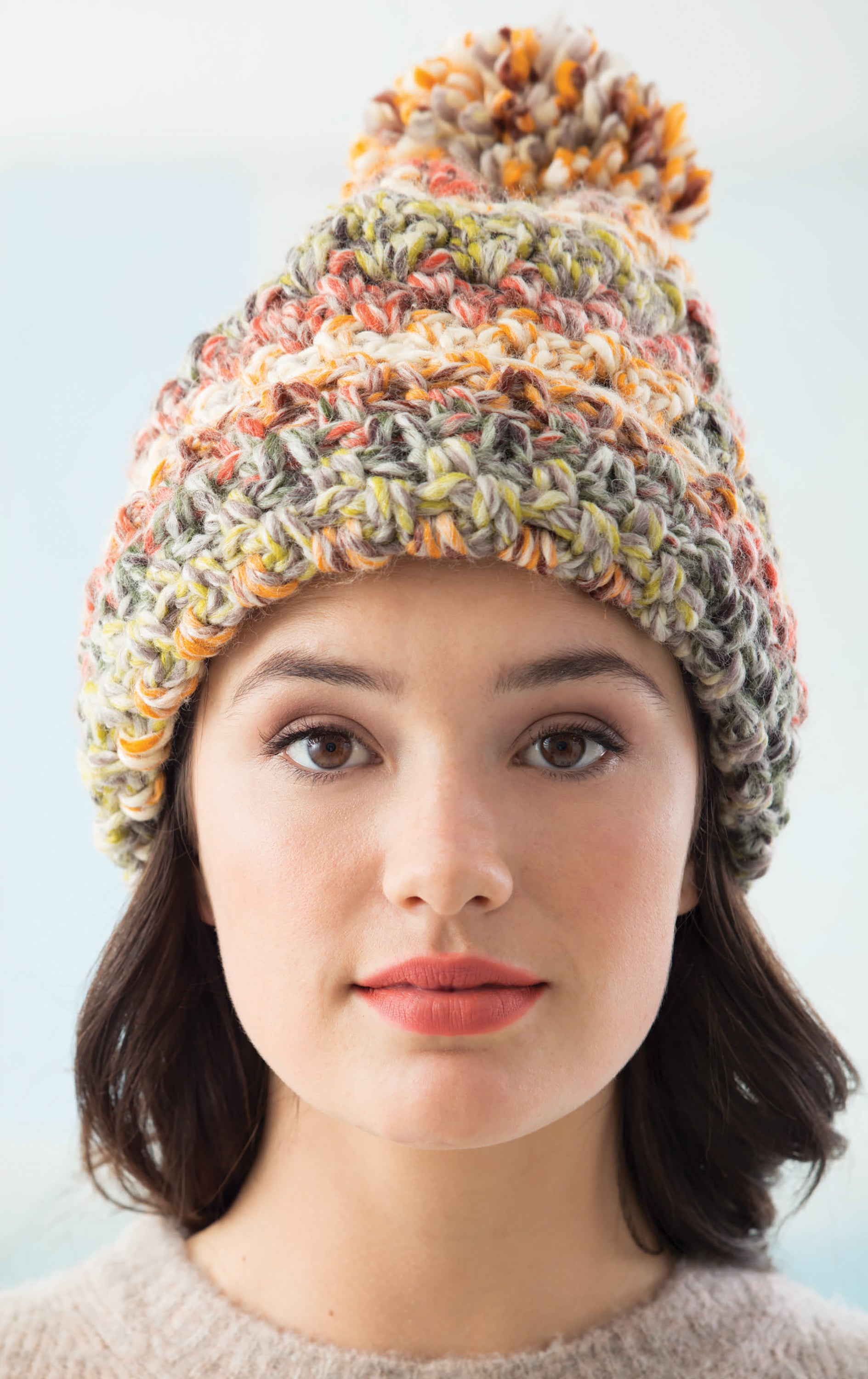 A person with short dark hair models a chunky, multicolored knit hat with pom-pom from Leisure Arts' "All Your Favorite Hats," gazing neutrally at the camera against a softly blurred background—ideal inspiration for new hat patterns.