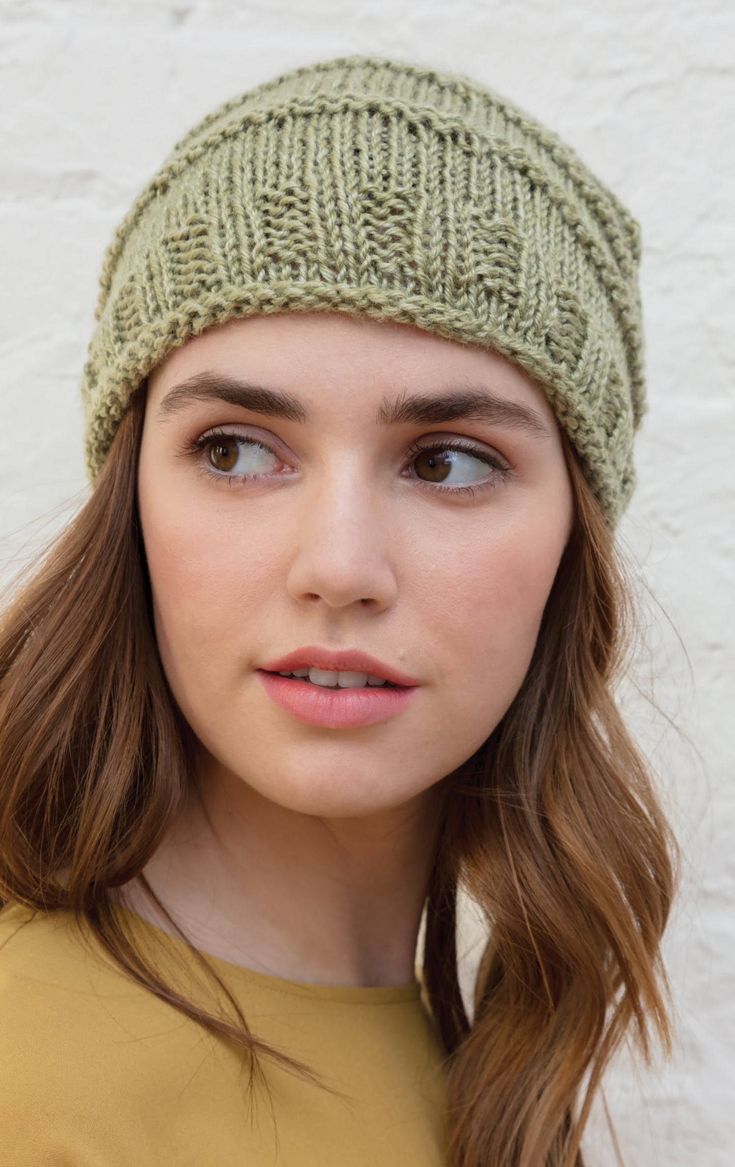 A young woman with long brown hair wears a light green hat from Leisure Arts' "All Your Favorite Hats" collection, pairing it with a mustard yellow top and posing against a white textured background.