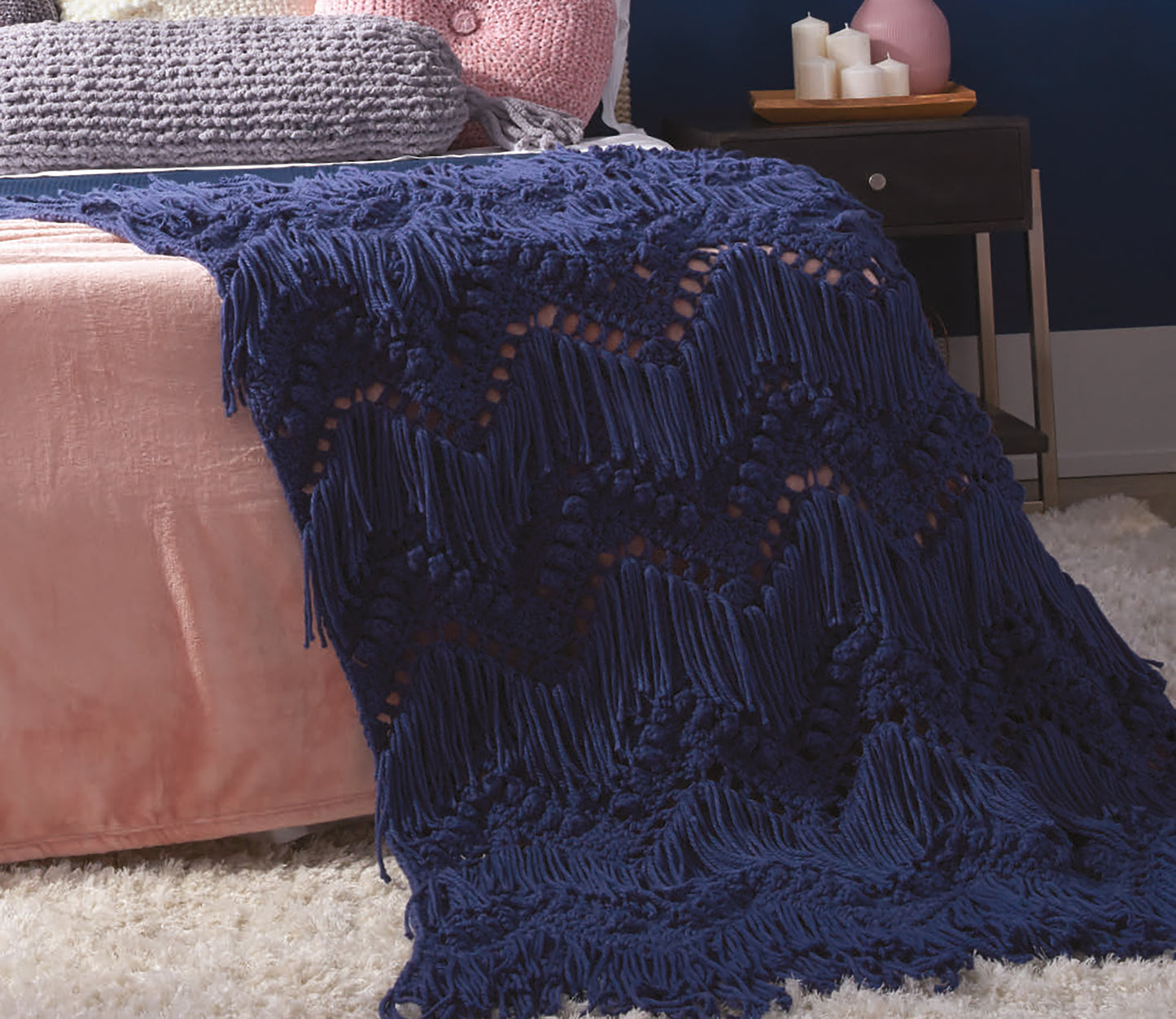 A pink bed is draped with a navy blue chevron macramé blanket made using bulky yarn and the Daydream Pattern Collection Digital Download from Leisure Arts. Nearby, a nightstand with candles sits on a plush cream rug.