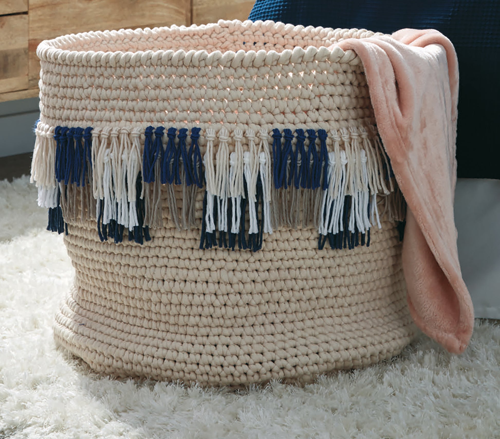 A soft pink blanket, created with bulky yarn and the Daydream Pattern Collection Digital Download by Leisure Arts, is draped over a large beige woven basket decorated with blue, white, and gray tassels on a fluffy white rug.