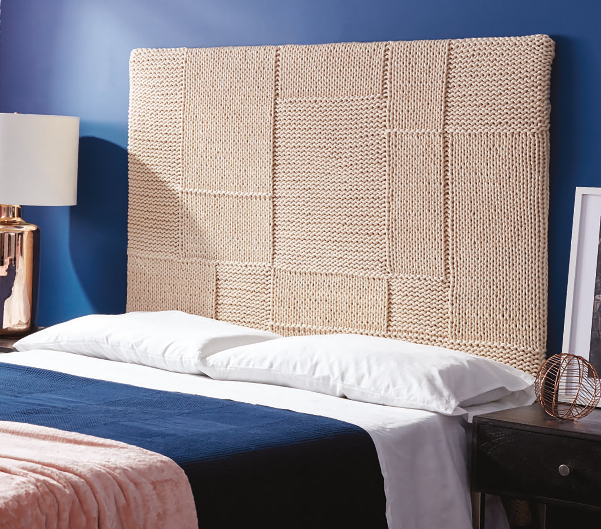 A bed with white pillows, a navy blanket, and a pink throw from the Daydream Pattern Collection by Leisure Arts sits before a beige patchwork headboard and dark blue wall, with a lamp and framed art on the nightstand.