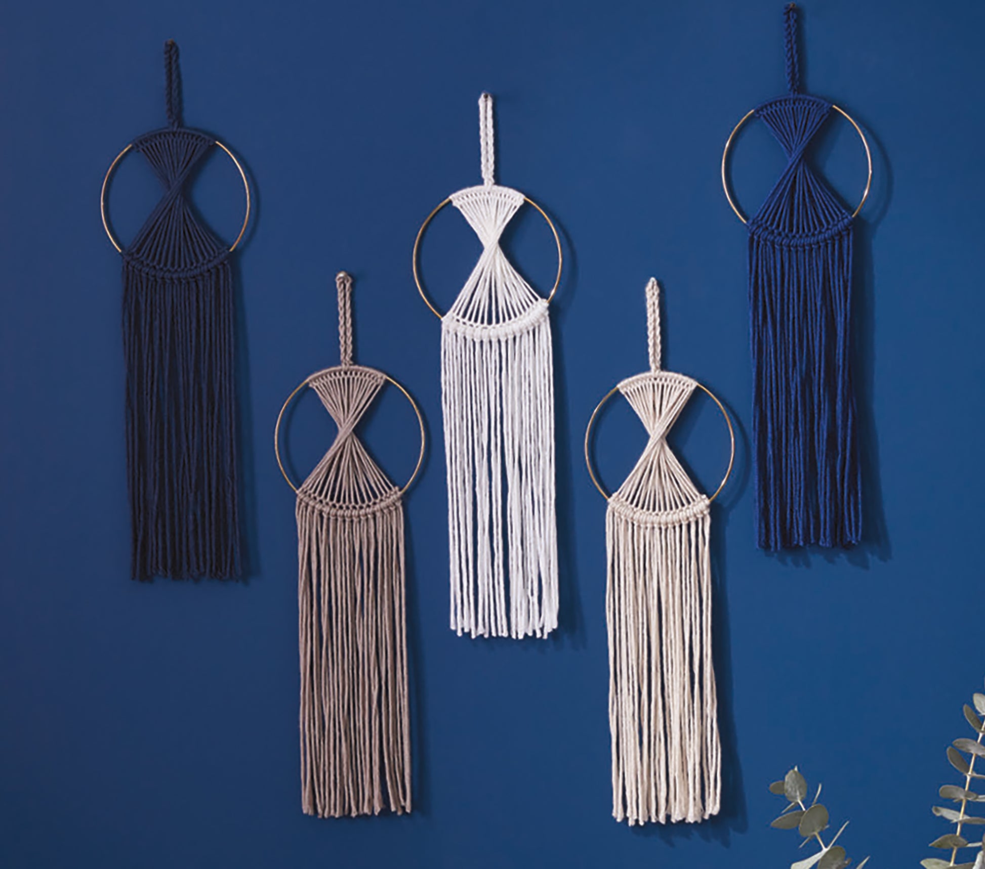 Five circular macramé wall hangings, inspired by crochet and knit with bulky yarn, adorn a blue wall. Created from the Daydream Pattern Collection Digital Download by Leisure Arts, each piece features metal rings and fringe; leaves peek nearby.