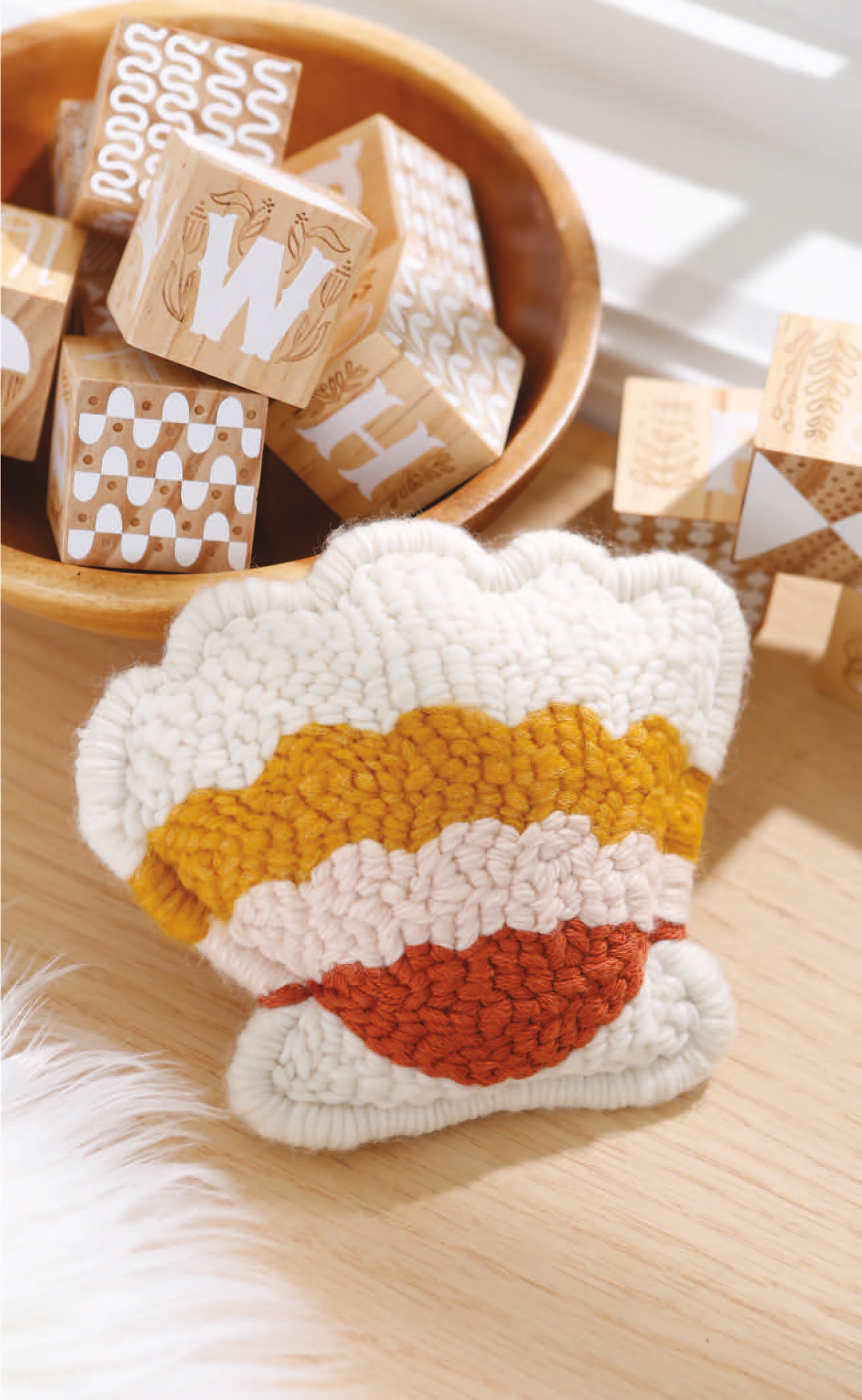 A copy of "Baby Punch Needle—Step-by-Step Instructions for 13 Projects" by Leisure Arts rests on a wooden surface near wooden alphabet blocks, highlighted by sunlight filtering in from the side.