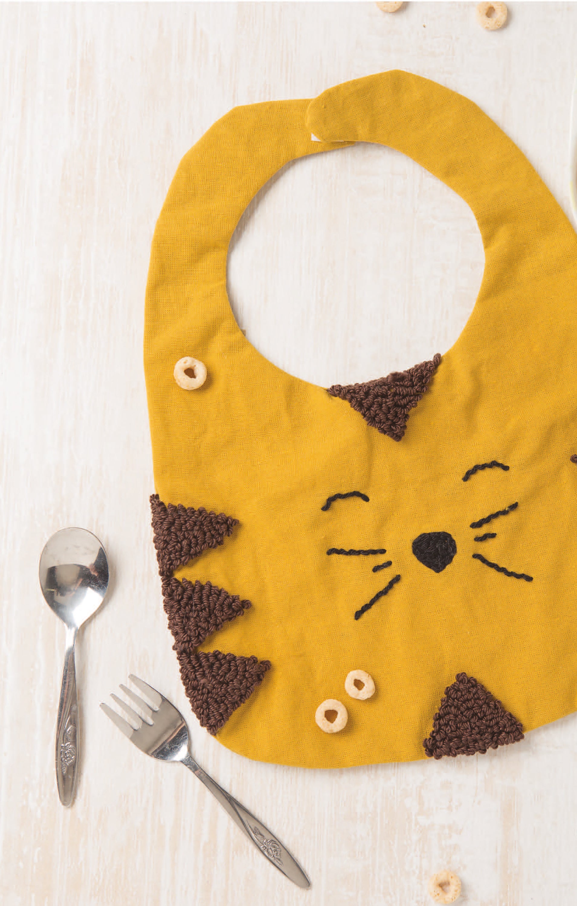 The book "Baby Punch Needle—Step-by-Step Instructions for 13 Projects" by Leisure Arts is displayed on a light wooden surface, surrounded by trendy punch needle designs. A spoon and fork lie to the left, with colorful cereal pieces scattered nearby.