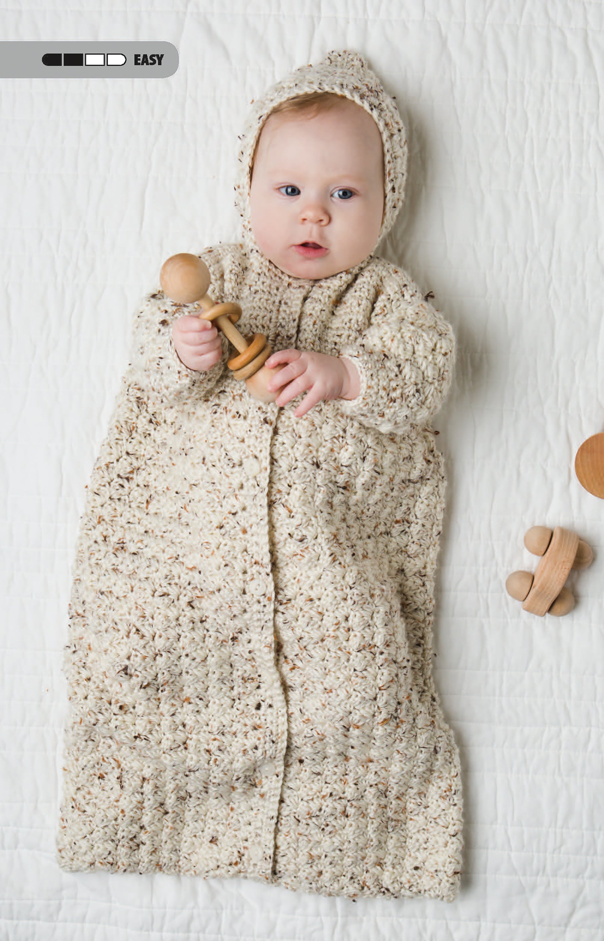 A baby in a cream, textured Crochet Tiny Tot Accessories sleep sack and matching bonnet by Leisure Arts lies on a white quilt, holding a wooden rattle. Wooden toys sit nearby, with an easy skill level icon for beginner-friendly crochet baby patterns.