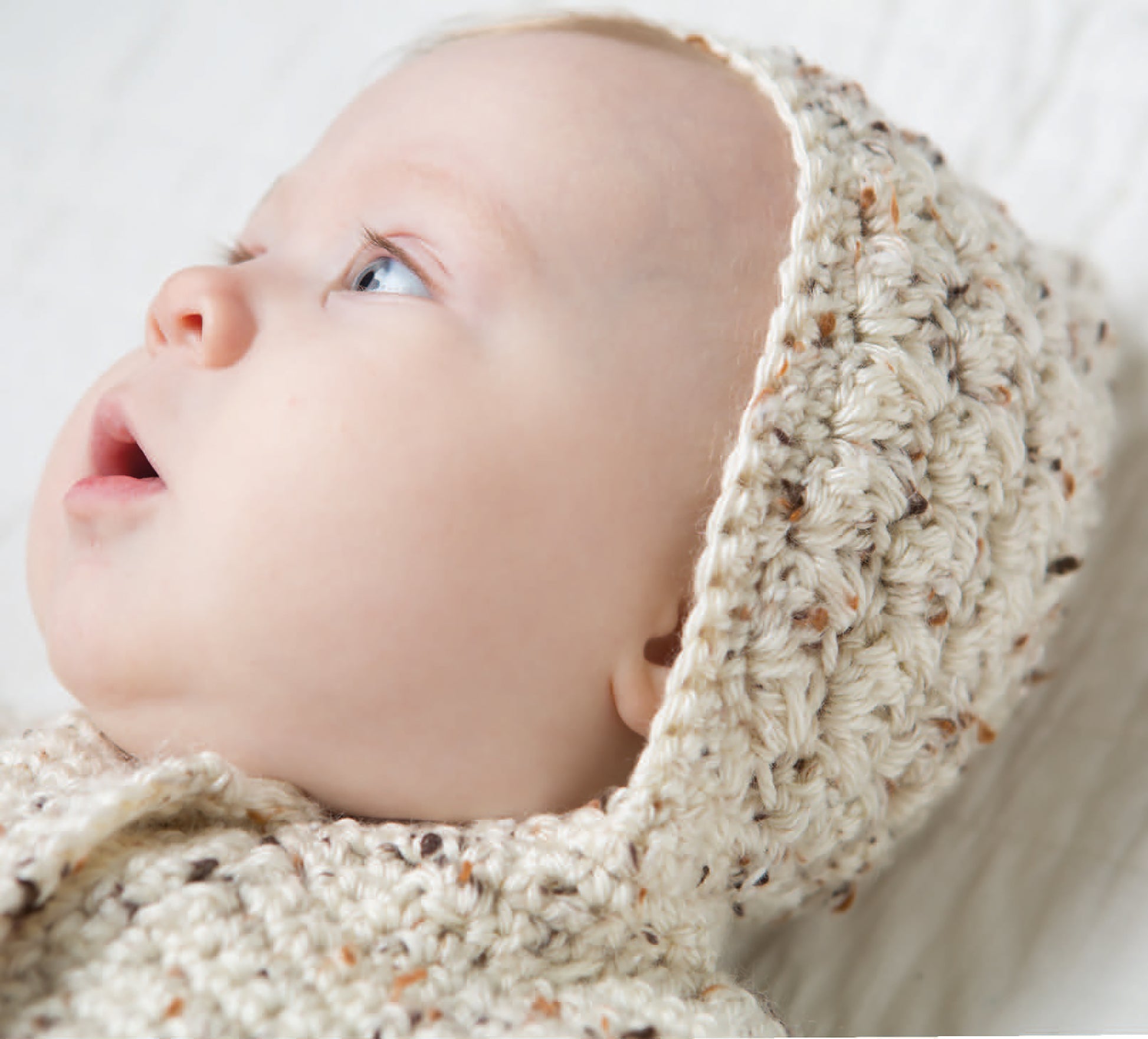 A fair-skinned, blue-eyed baby gazes upward while bundled in a cream crocheted hooded outfit, a sweet example from Leisure Arts' Crochet Tiny Tot Accessories—perfect for easy and adorable baby crochet inspiration.