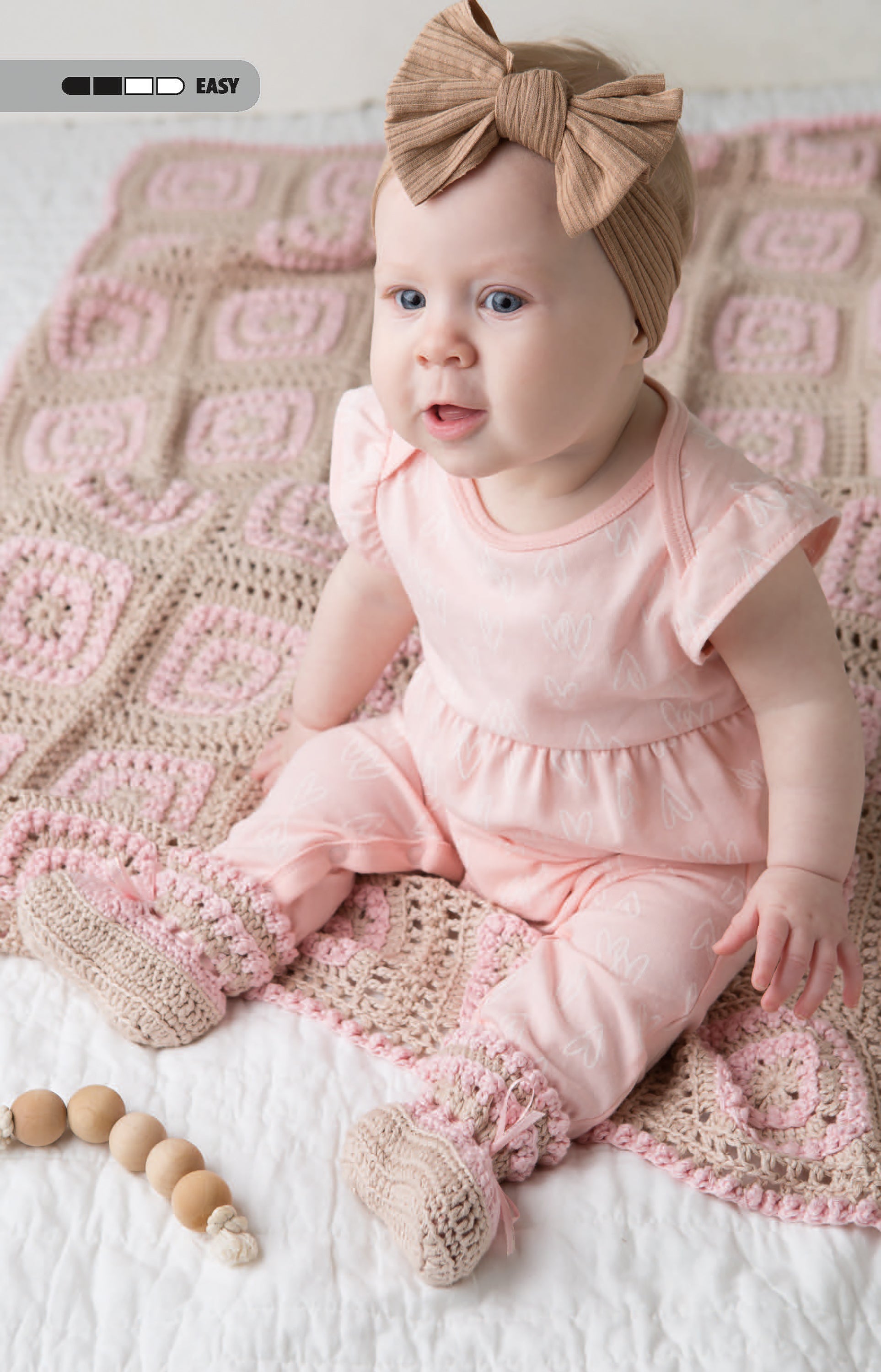 A baby in pink and a brown bow headband sits on a cozy blanket with a wooden bead toy—perfect inspiration from Leisure Arts' Crochet Tiny Tot Accessories for easy baby crochet projects.