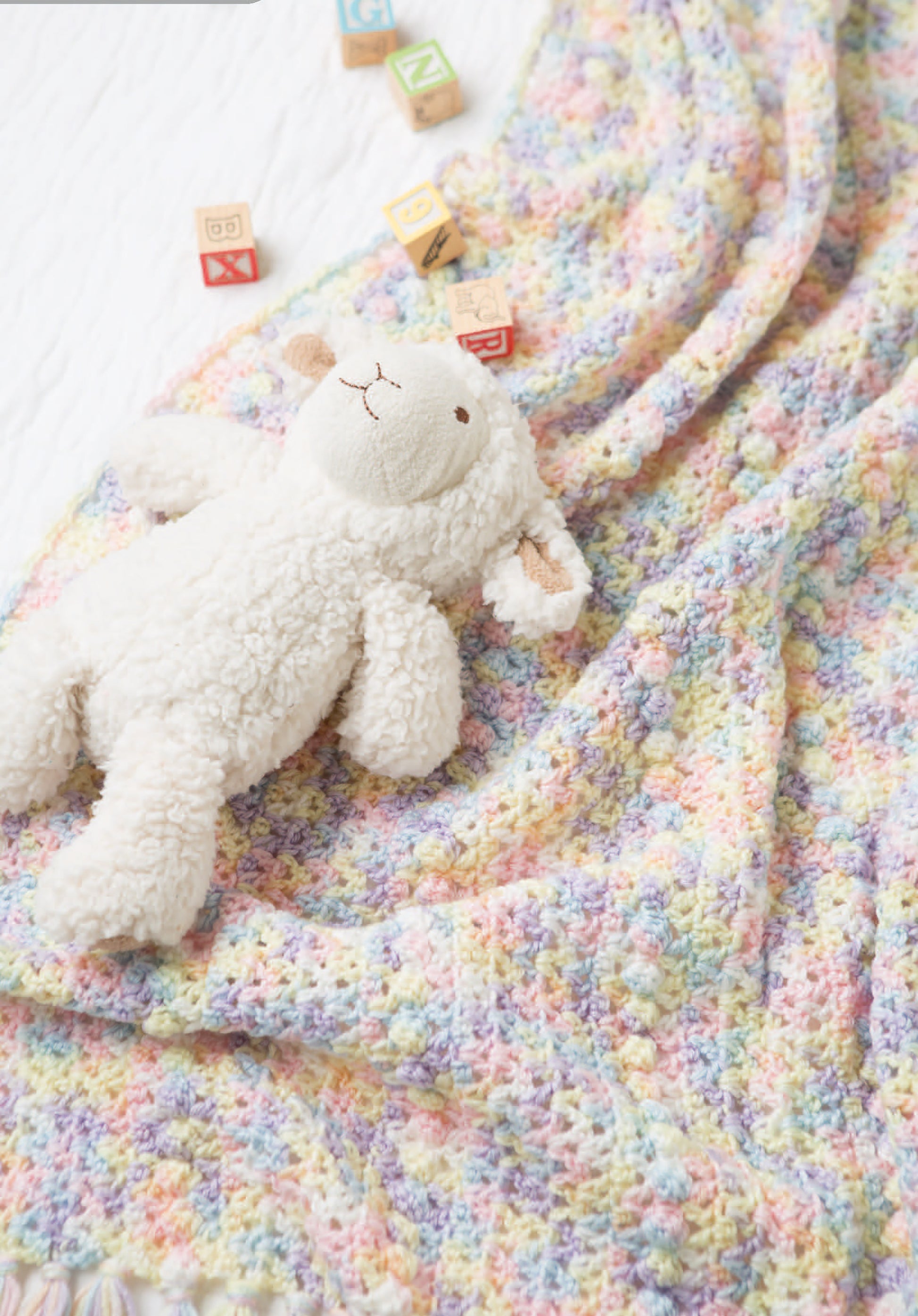 A cuddly stuffed lamb rests on a pastel newborn crochet blanket, accented by wooden alphabet blocks—just like the charming designs you’ll find in Leisure Arts’ Crochet Tiny Tot Accessories pattern book.