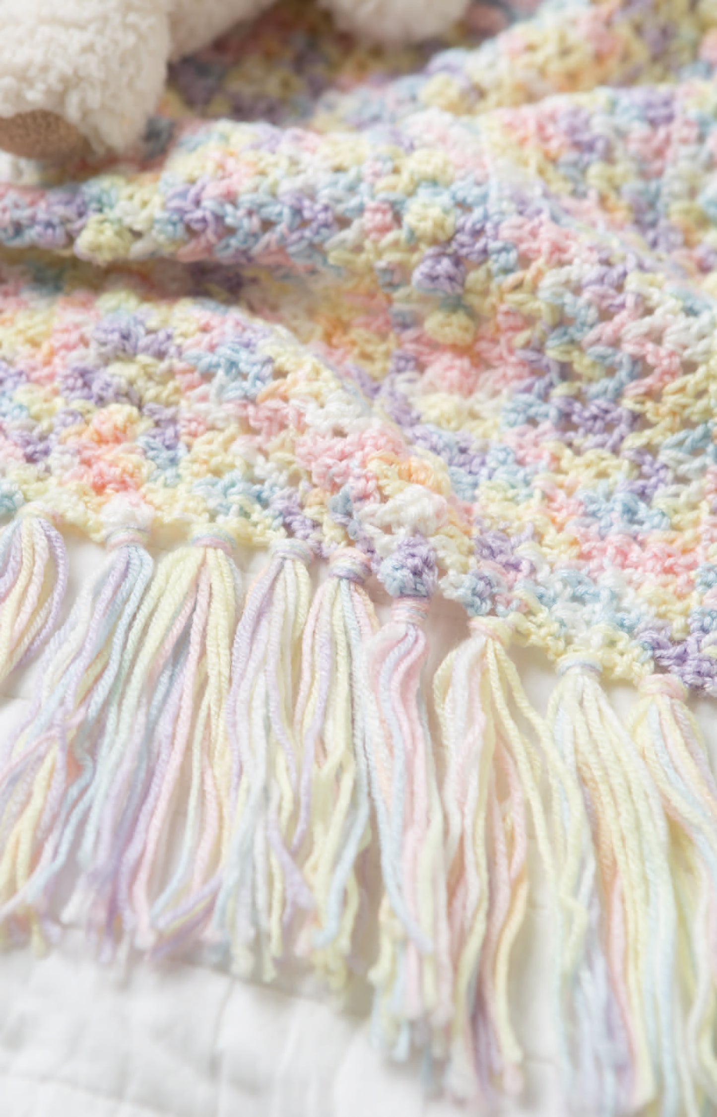 A close-up of a pastel-colored newborn crochet blanket with fringe edges from Leisure Arts’ Crochet Tiny Tot Accessories, featuring soft yellow, pink, blue, and purple—a perfect choice for easy baby crochet projects.