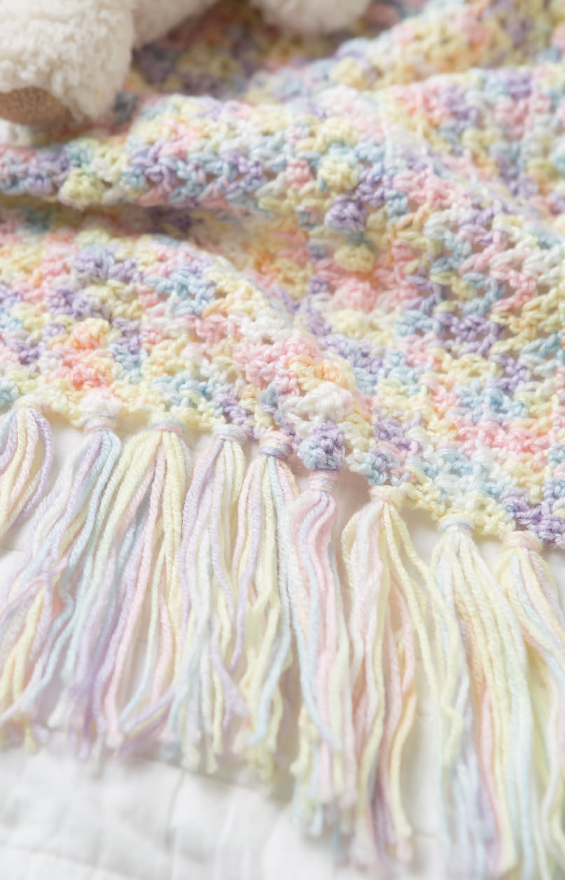 A close-up of a pastel-colored newborn crochet blanket with fringe edges from Leisure Arts’ Crochet Tiny Tot Accessories, featuring soft yellow, pink, blue, and purple—a perfect choice for easy baby crochet projects.