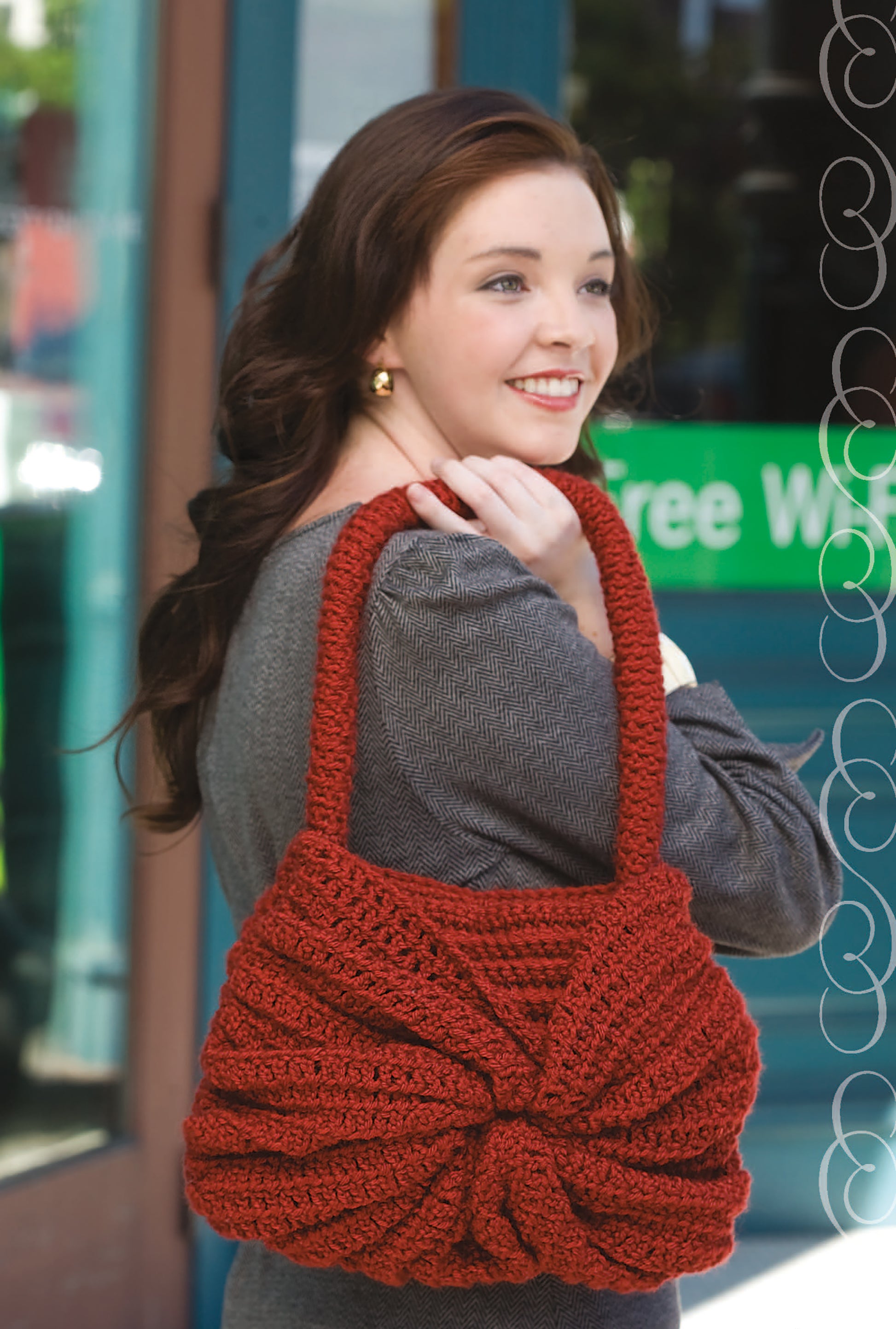 A smiling woman with long brown hair showcases the Must Have Handbags Digital Download by Leisure Arts, featuring unique crochet designs, as she stands outside near a glass door holding a large red textured handbag.