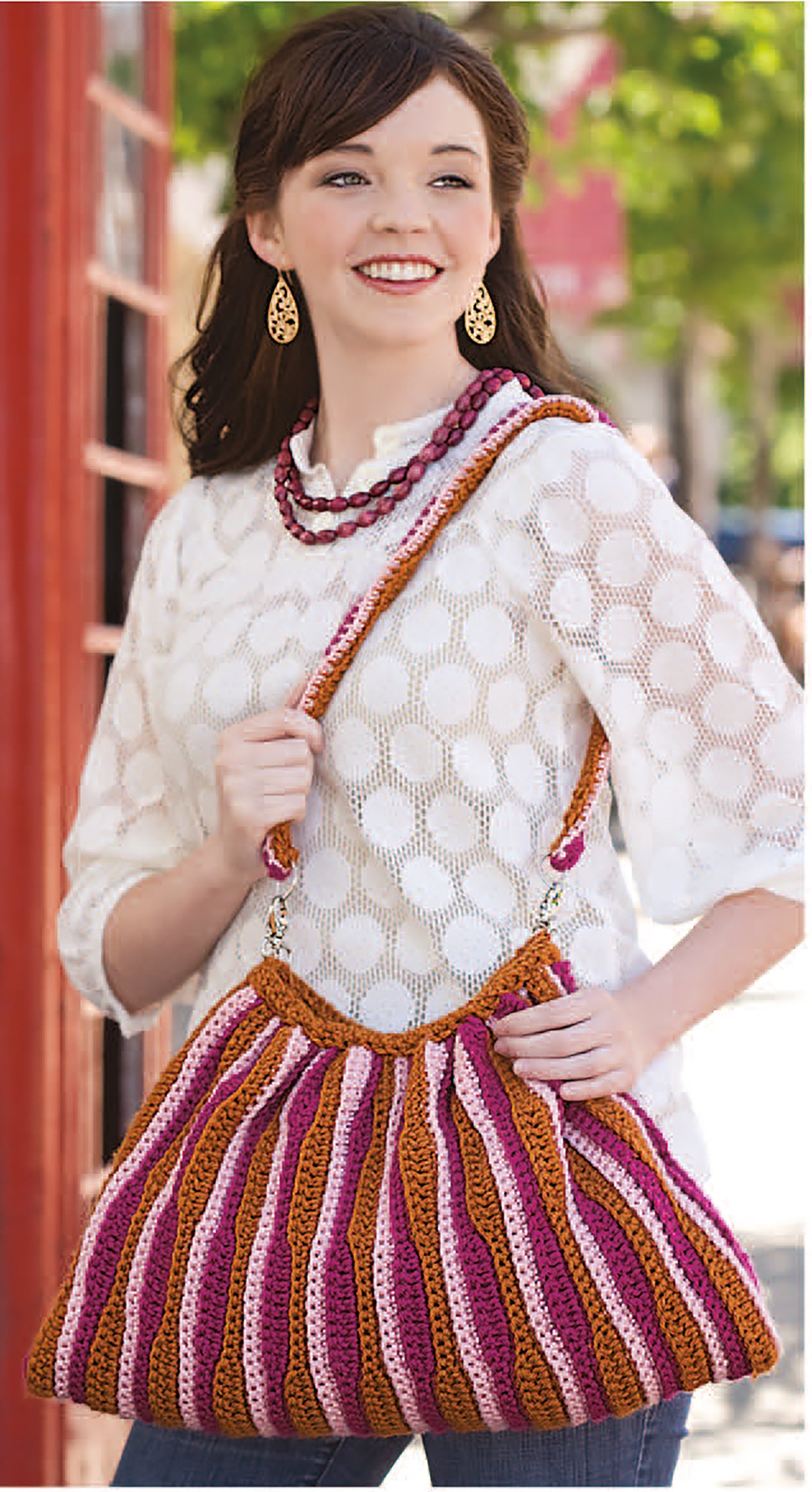 A smiling woman outdoors holds a large, colorful crochet bag over her shoulder—showcasing unique designs from the Must Have Handbags Digital Download by Leisure Arts. Gold earrings and a white patterned top complete her stylish look.