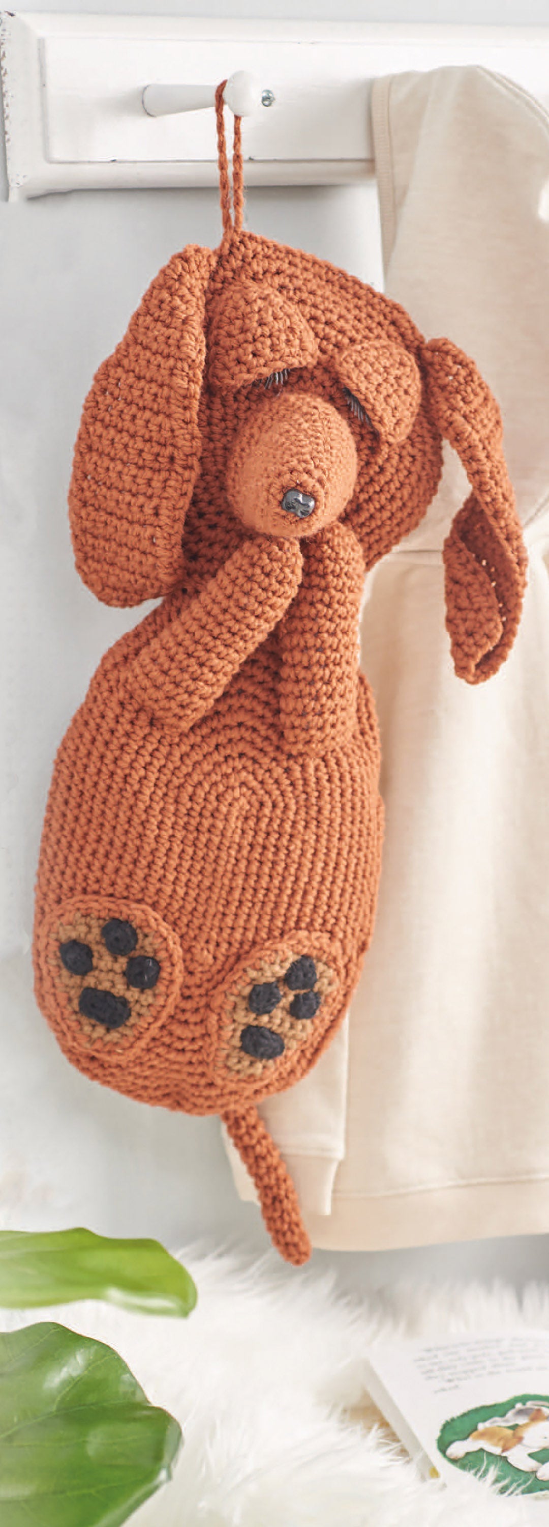 The Cuddle Buddies Crochet bag by Leisure Arts—a brown, dog-shaped design with floppy ears and black paw prints—hangs on a white hook beside a cream garment, perfect for crochet animal fans.