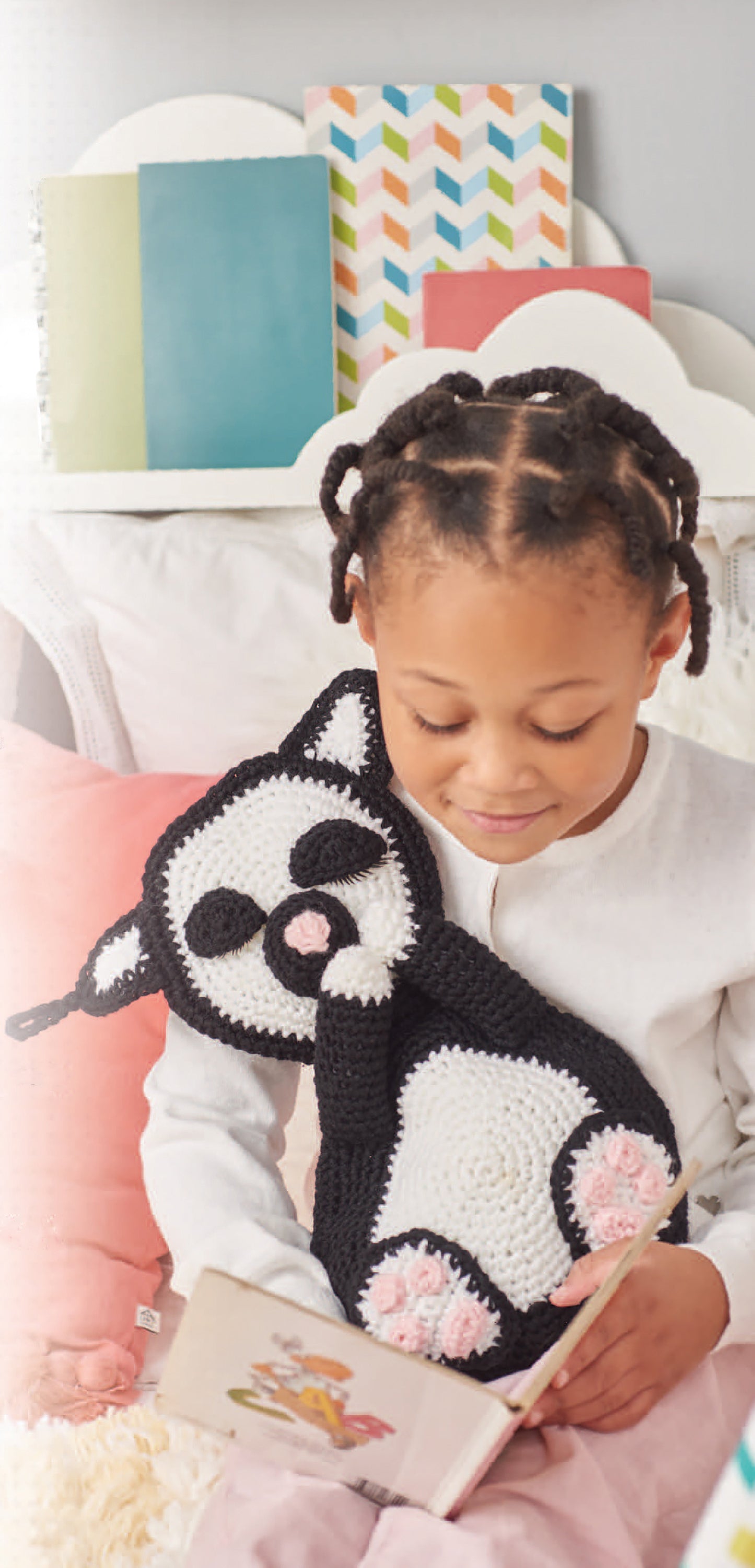 A young girl with braided hair sits on a bed, holding a Leisure Arts Cuddle Buddies Crochet black and white cat plush while reading. Colorful notebooks and pillows can be seen in the background.