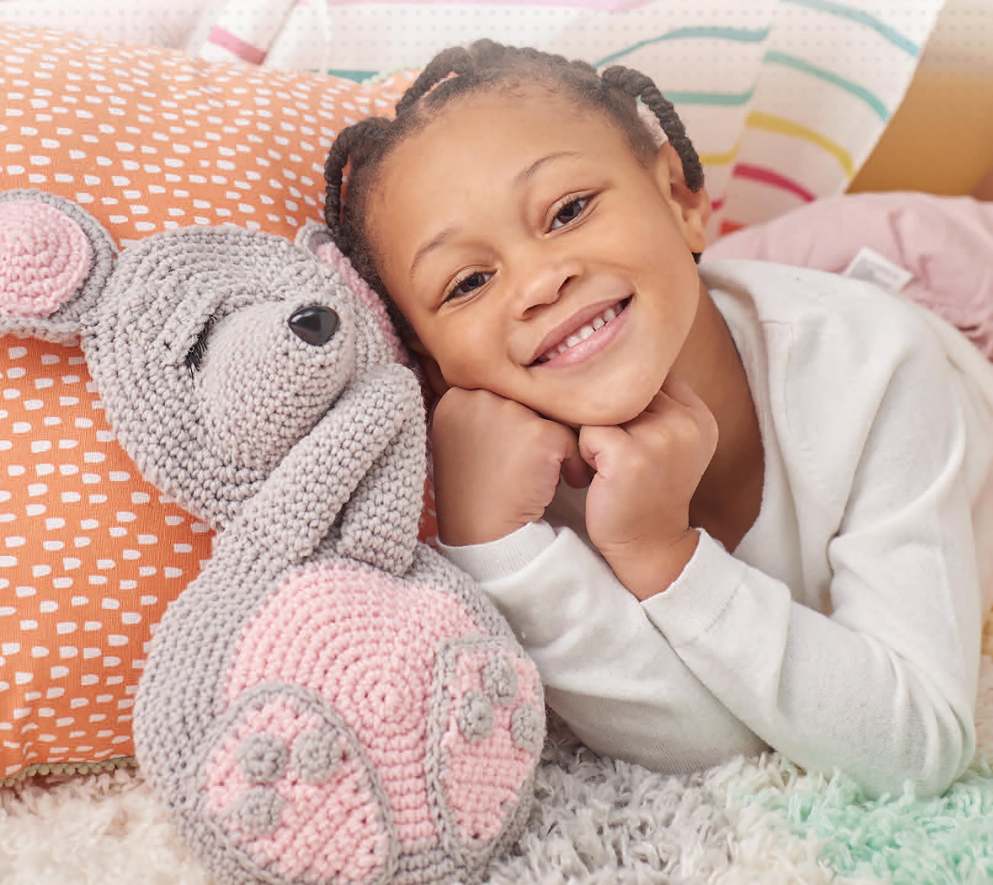 A young child with braided hair smiles while lying on a bed, resting their chin on their hands next to a crocheted mouse made with Cuddle Buddies Crochet by Leisure Arts. Colorful pillows and patterned blankets add to the cozy scene.