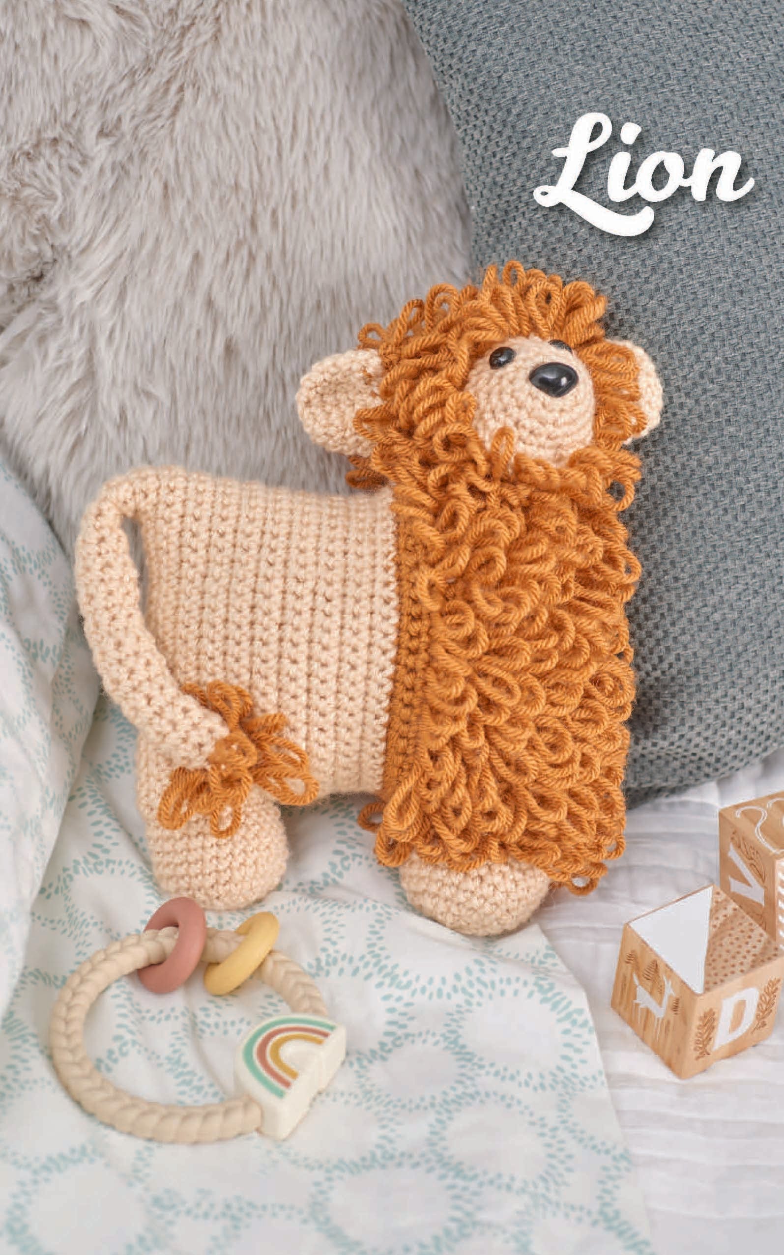 A Car Seat Cuties crocheted lion toy from Leisure Arts with a curly orange mane rests on a bed with cushions, baby blocks, and a teething ring. Ideal for beginner crochet projects or fans of crochet animal patterns.