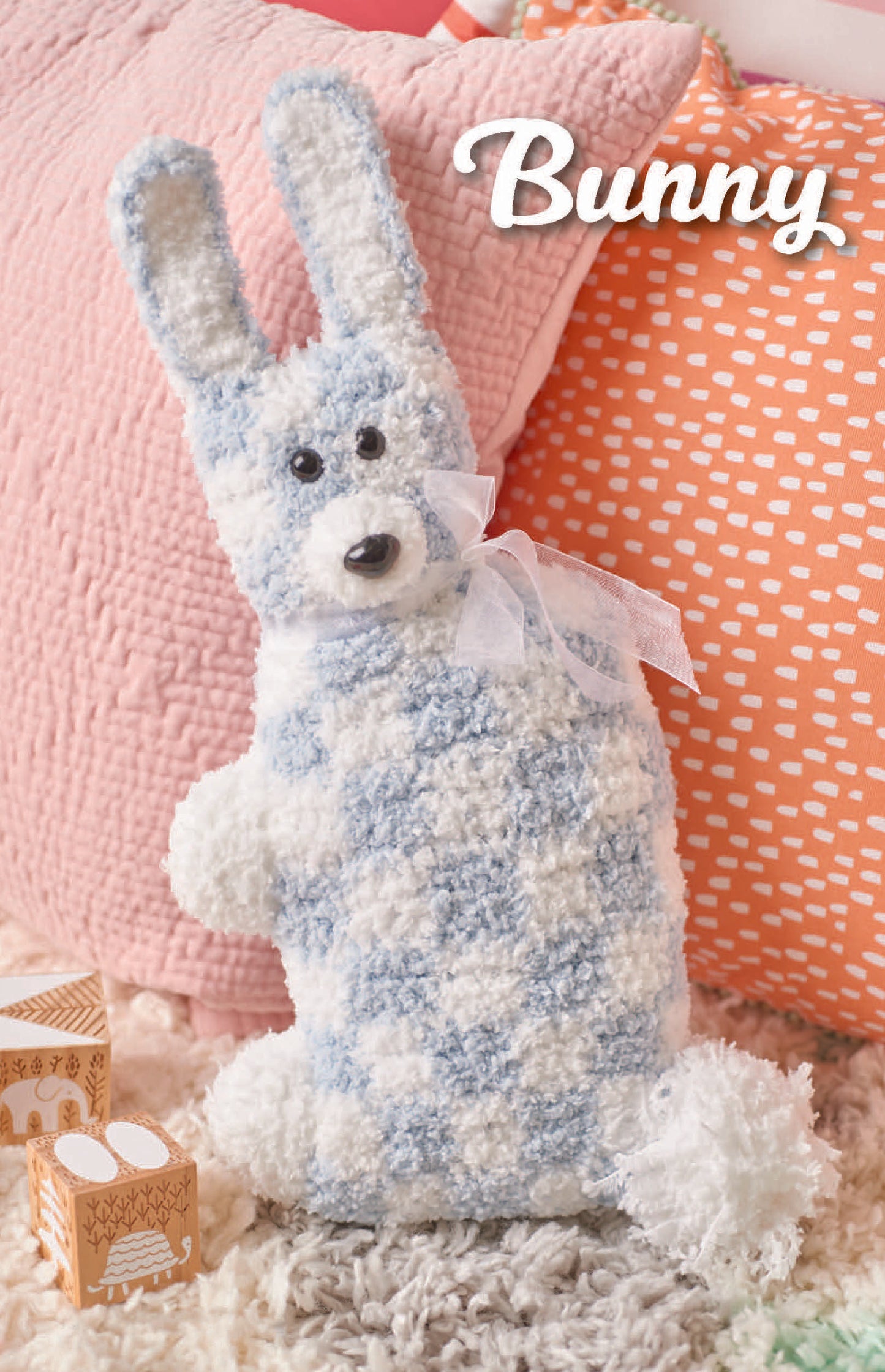 Car Seat Cuties by Leisure Arts: a soft blue-and-white checkered plush bunny with a sheer white ribbon sits on a textured rug before peach and orange pillows—sweet inspiration for beginner crochet projects. "Bunny" appears in white at the top right.