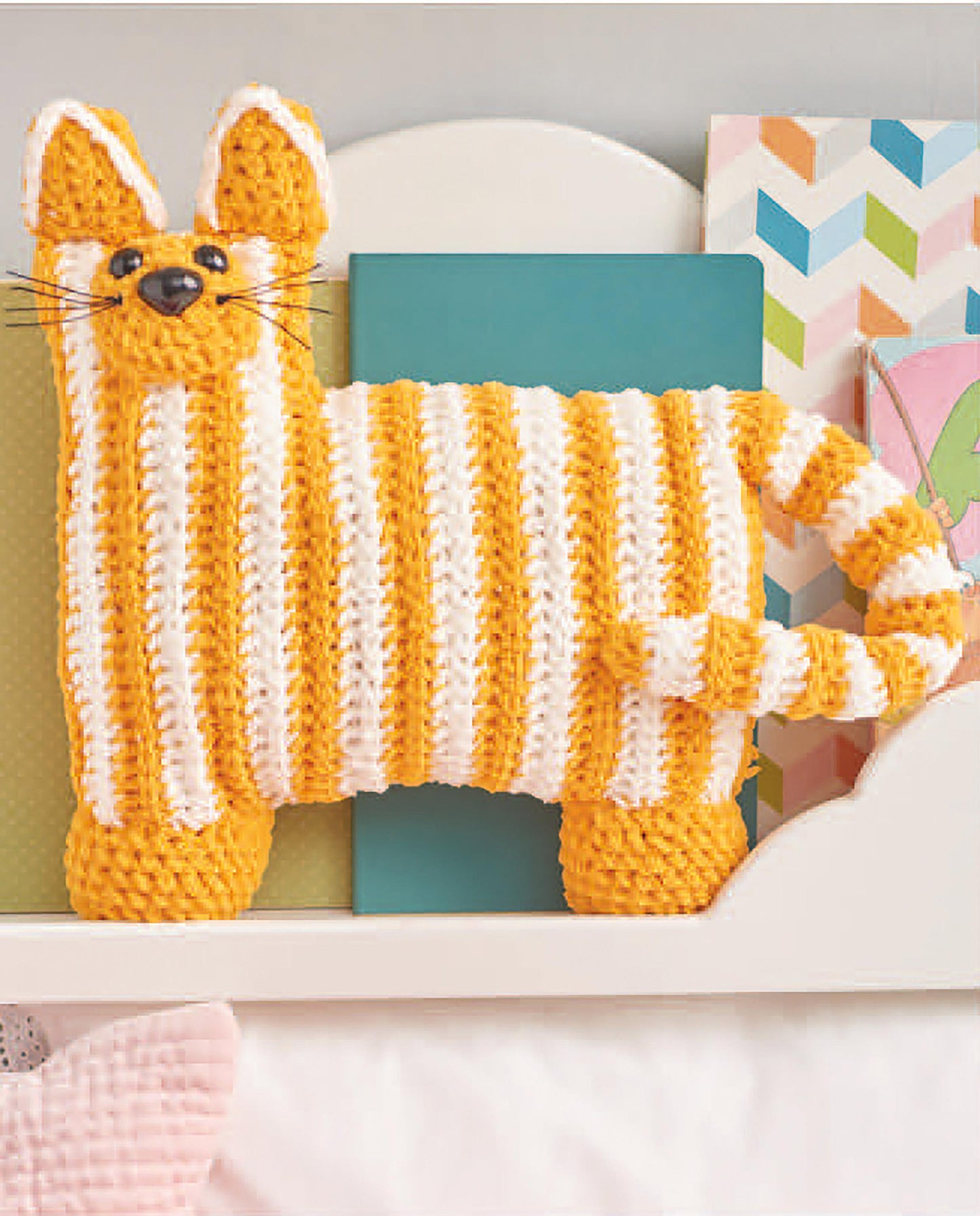 The Car Seat Cuties plush cat from Leisure Arts, with orange and white stripes, pointy ears, a striped tail, black whiskers, and a stitched smile, sits in front of colorful books and décor—ideal for crochet animal pattern fans.