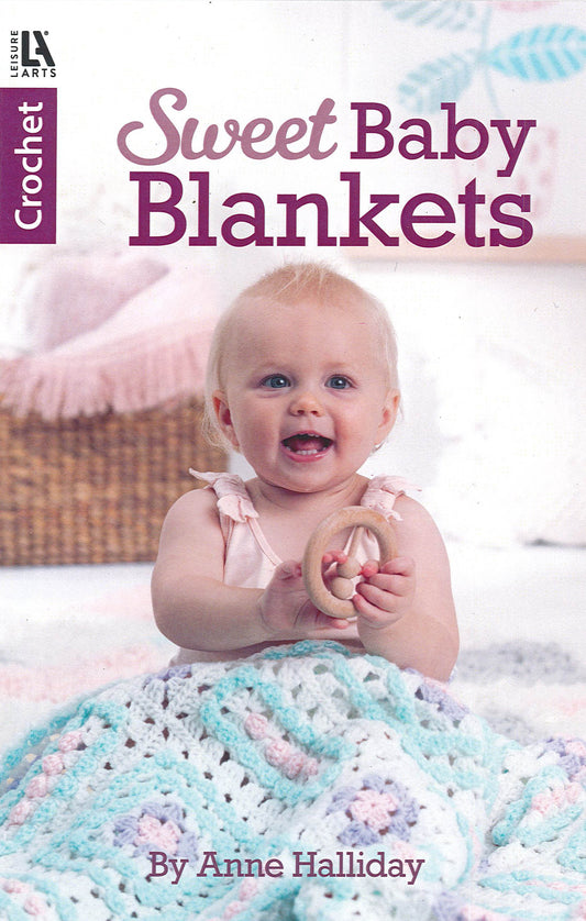 A smiling baby sits on a bed holding a wooden teether, partly covered by a pastel crocheted blanket. The Sweet Baby Blankets Digital Download from Leisure Arts offers beginner crochet patterns, ideal for thoughtful handmade gifts.