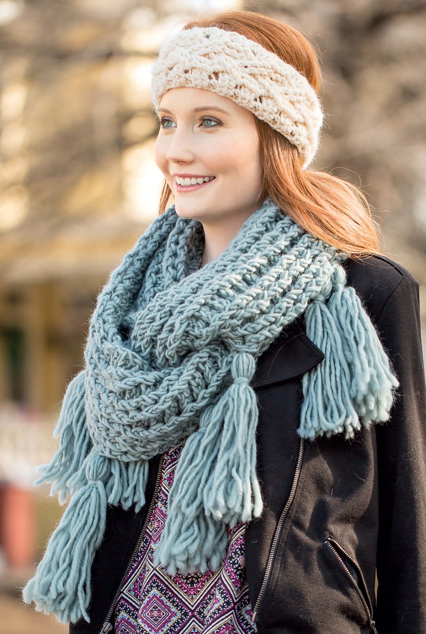 A woman with red hair smiles outdoors, wearing cozy crochet accessories inspired by Leisure Arts’ Big Book of Crochet Accessories—a chunky cream knit headband and a large light blue tasseled scarf—embracing warmth in a softly blurred setting.