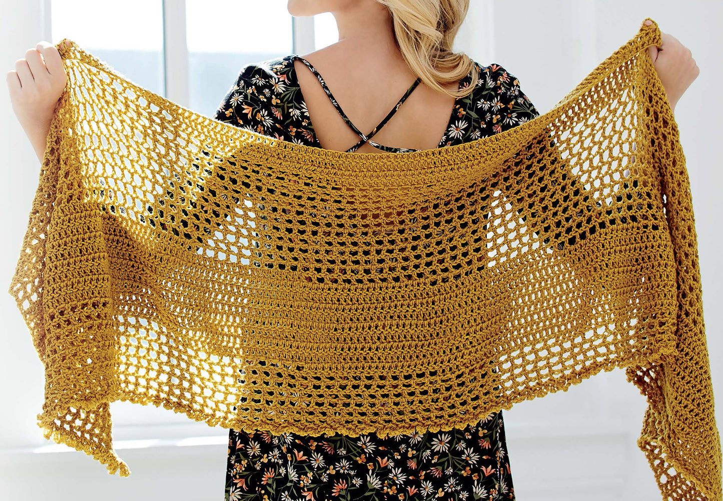 A person in a floral dress displays a wide, mustard yellow crocheted shawl with an airy pattern from the Big Book of Crochet Accessories by Leisure Arts, perfect for pairing with hats or scarves in front of a bright window.