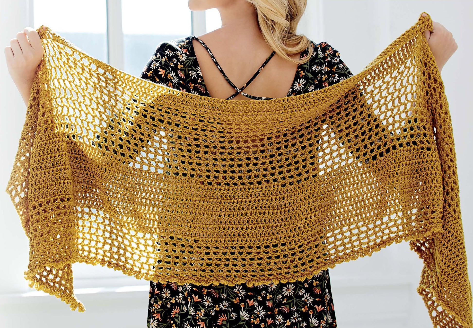 A person in a floral dress displays a wide, mustard yellow crocheted shawl with an airy pattern from the Big Book of Crochet Accessories by Leisure Arts, perfect for pairing with hats or scarves in front of a bright window.