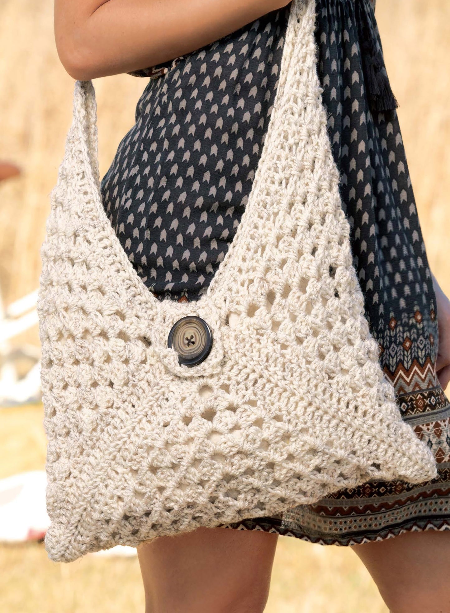 A person in a patterned dress carries a handmade off-white crochet shoulder bag with a central button, styled with hats and scarves. The accessories are featured in Leisure Arts’ Big Book of Crochet Accessories against an outdoor background.