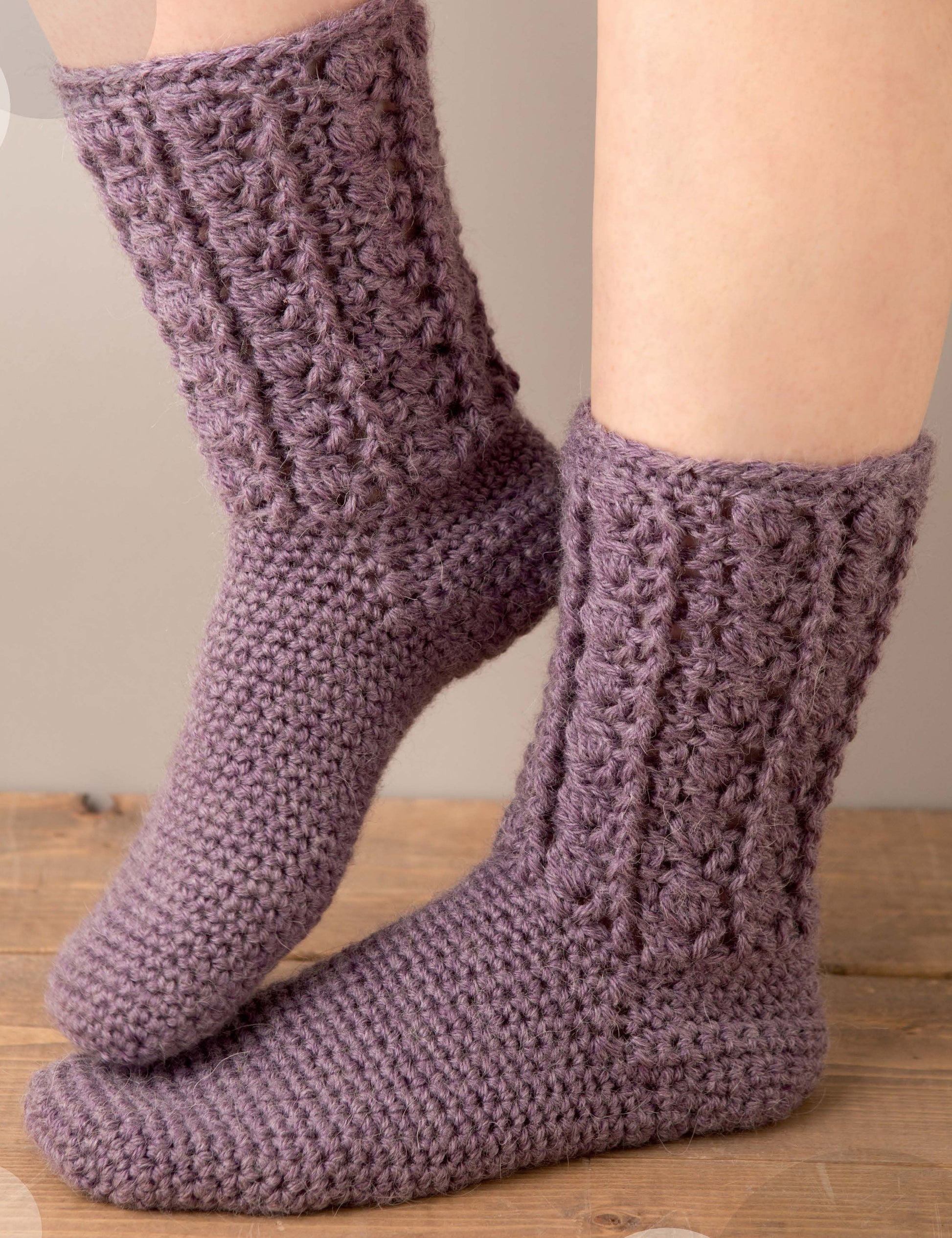 A cozy pair of textured purple socks is shown from the side on a wooden floor, featuring a chunky ribbed design—perfect for pairing with accessories from Leisure Arts' Big Book of Crochet Accessories.