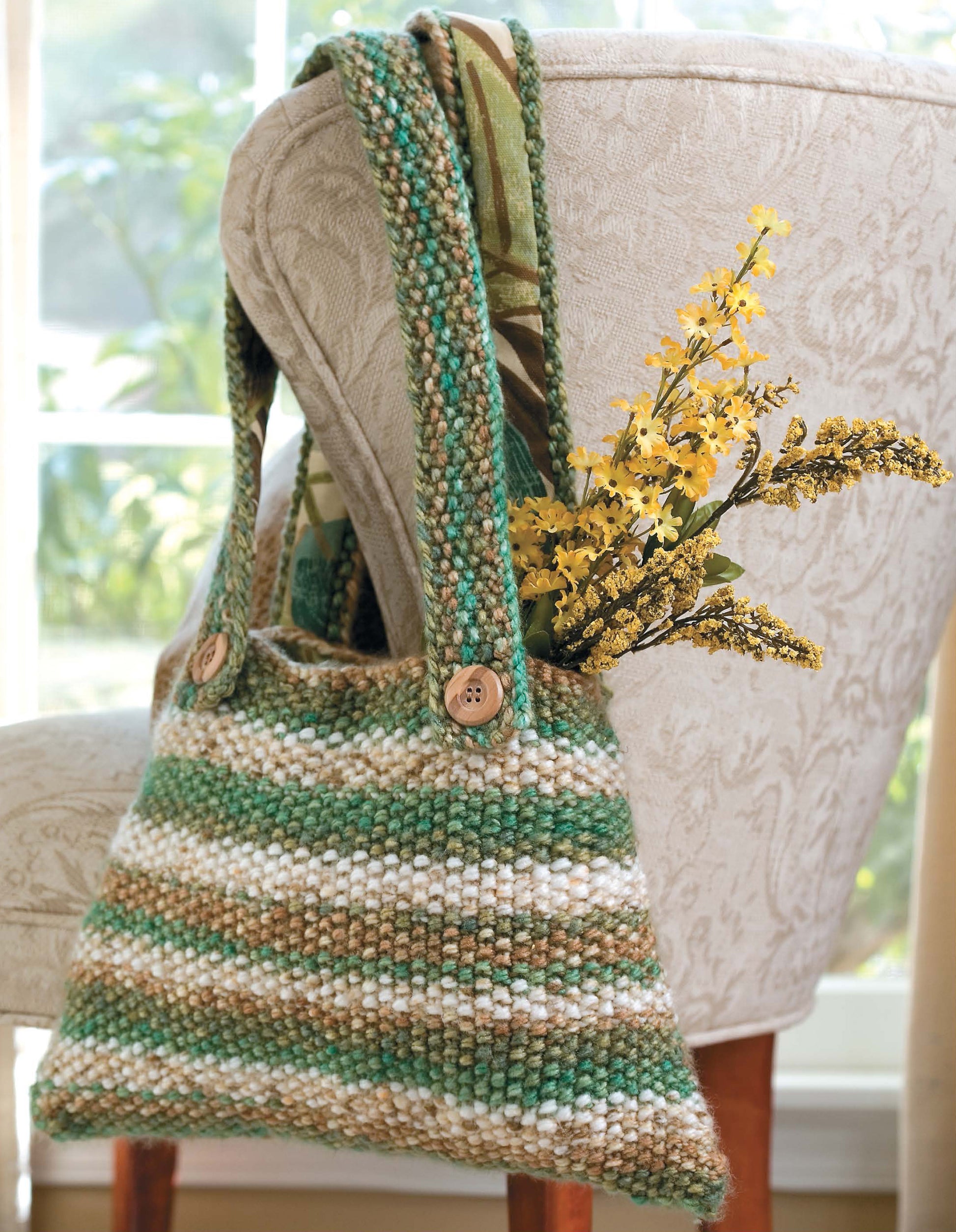 The Best of Knit Accessories Digital Download by Leisure Arts features hand-knitted designs like this green, beige, and white striped handbag with buttoned handles. Yellow flowers fill the bag as sunlight streams through a nearby window.