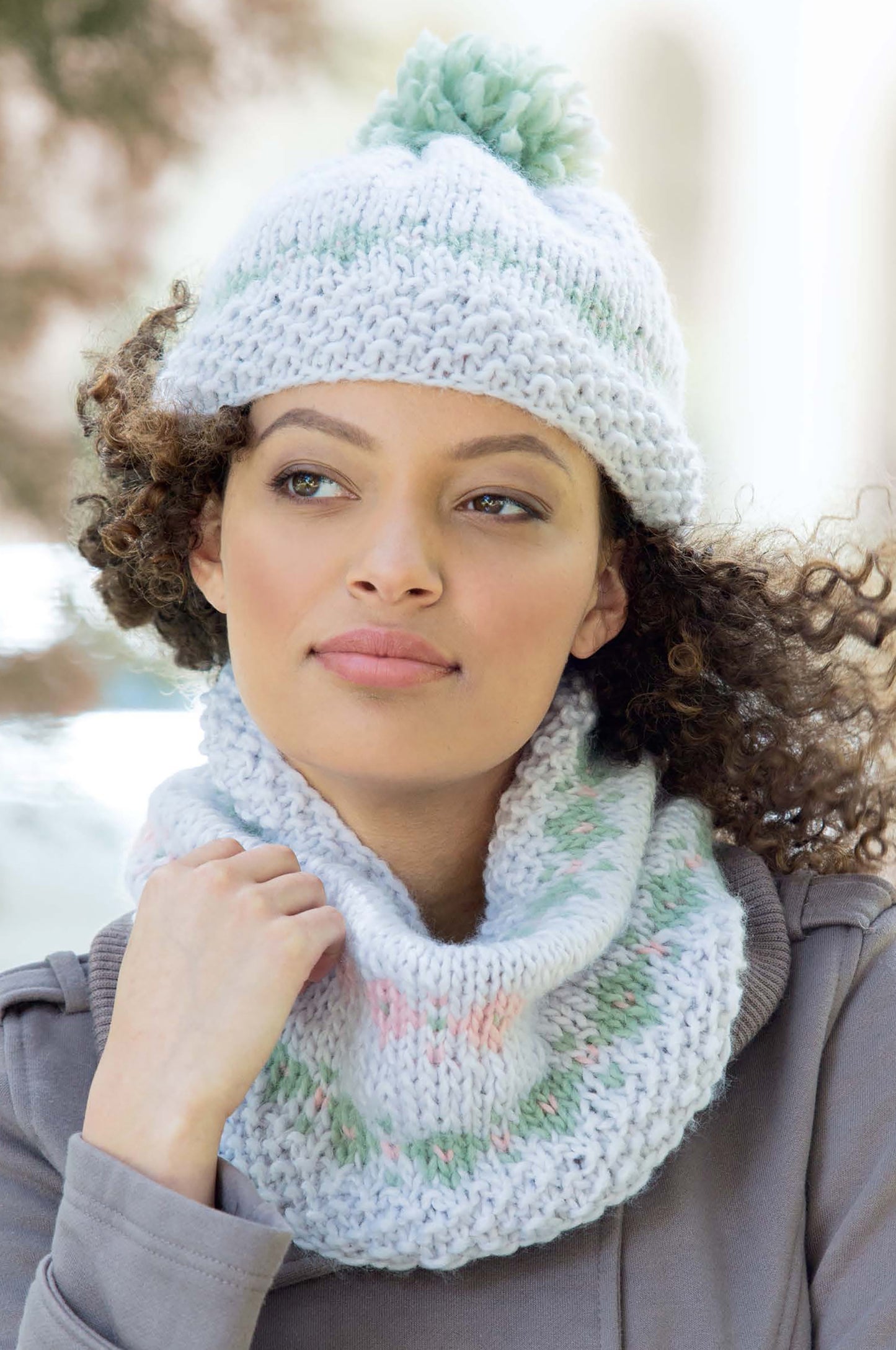 A curly-haired woman in a gray coat wears a hand-knit white hat with pom-pom and matching cowl—stylish projects from the Leisure Arts "Best of Knit Accessories" digital download—as she gazes calmly outdoors on a bright day.