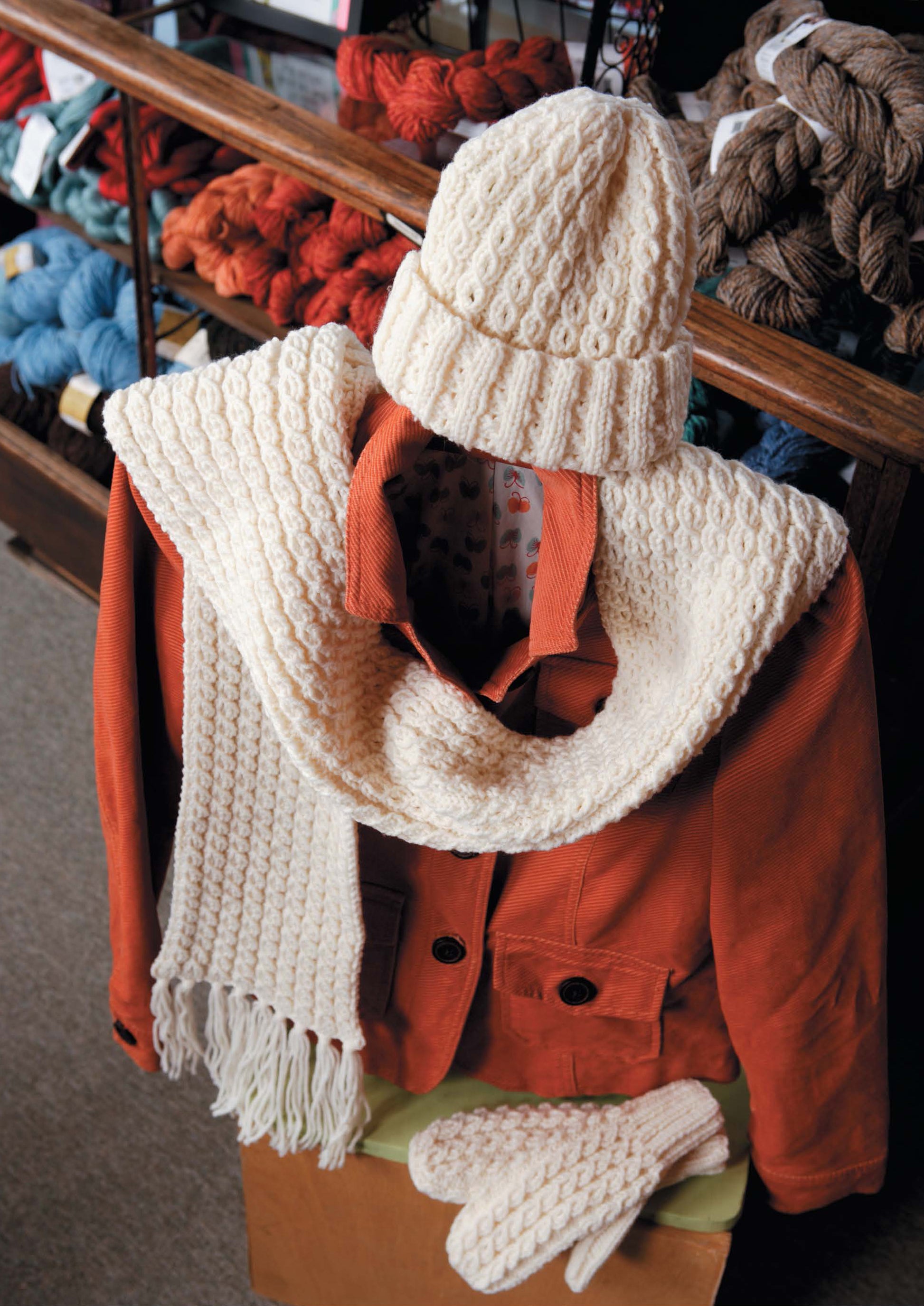 The Best of Knit Accessories Digital Download from Leisure Arts features hand-knitted cream accessories—hat, scarf, and mittens—shown on an orange jacket. Yarn skeins in blue, red, and brown are perfect for easy knitting projects.