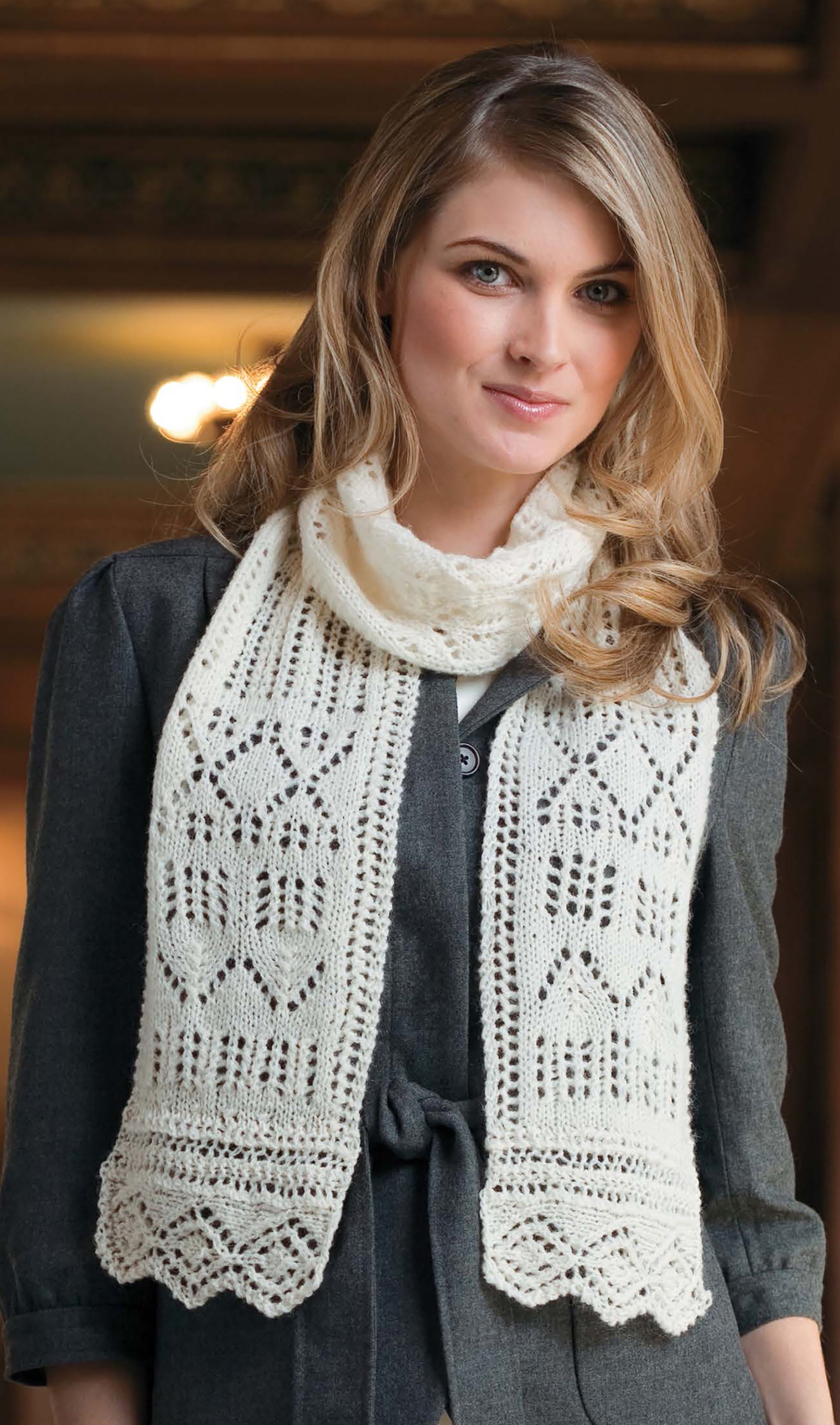A woman with long blonde hair wears a dark gray coat and a hand-knitted lace heart scarf—an inspiration from Leisure Arts’ Best of Knit Accessories Digital Download, perfect for easy scarf and mitt projects.