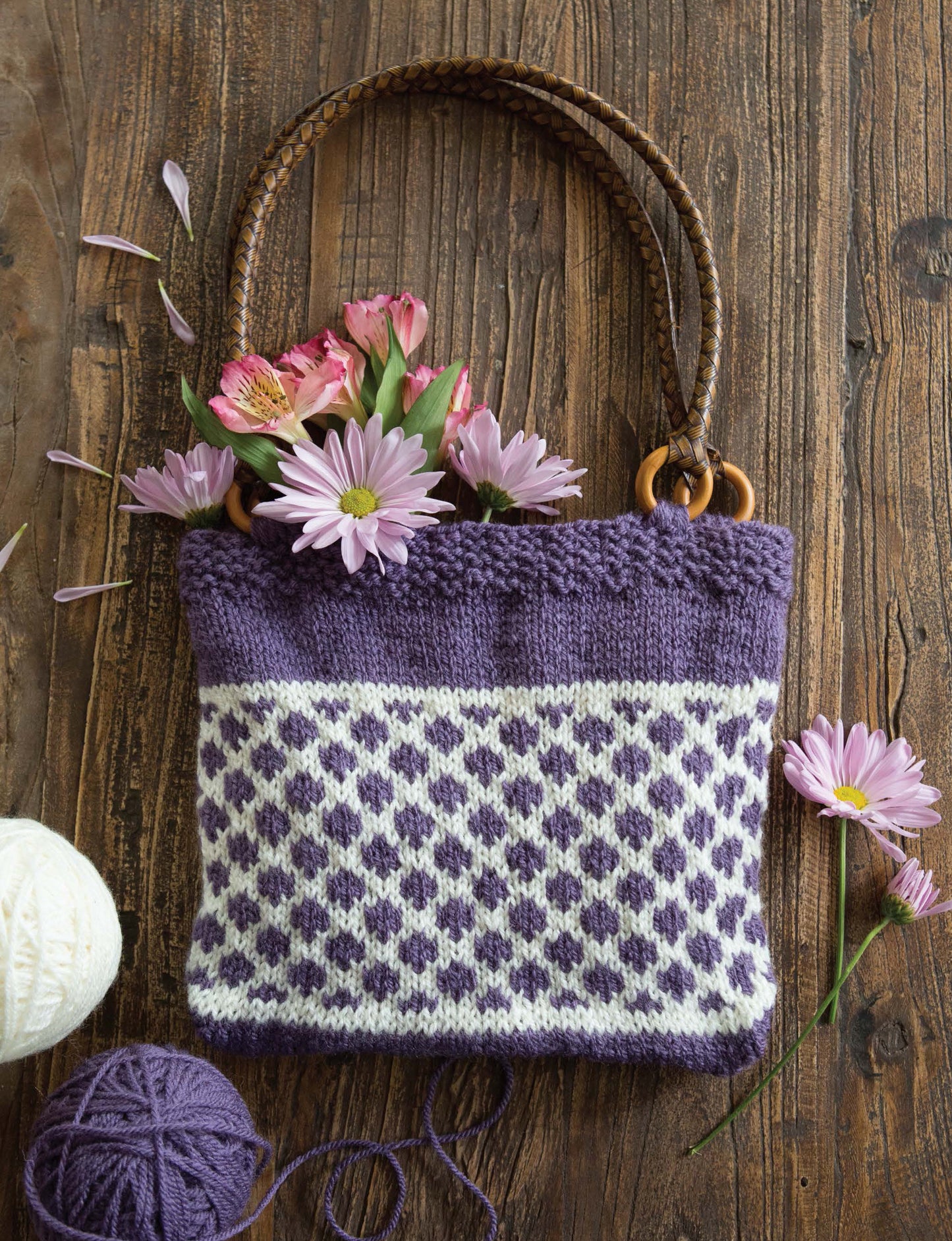 A rustic wooden surface holds a purple and white patterned knit bag with brown handles and wooden rings, overflowing with pink and lavender flowers—perfect inspiration for easy projects from Leisure Arts' Best of Knit Accessories Digital Download.