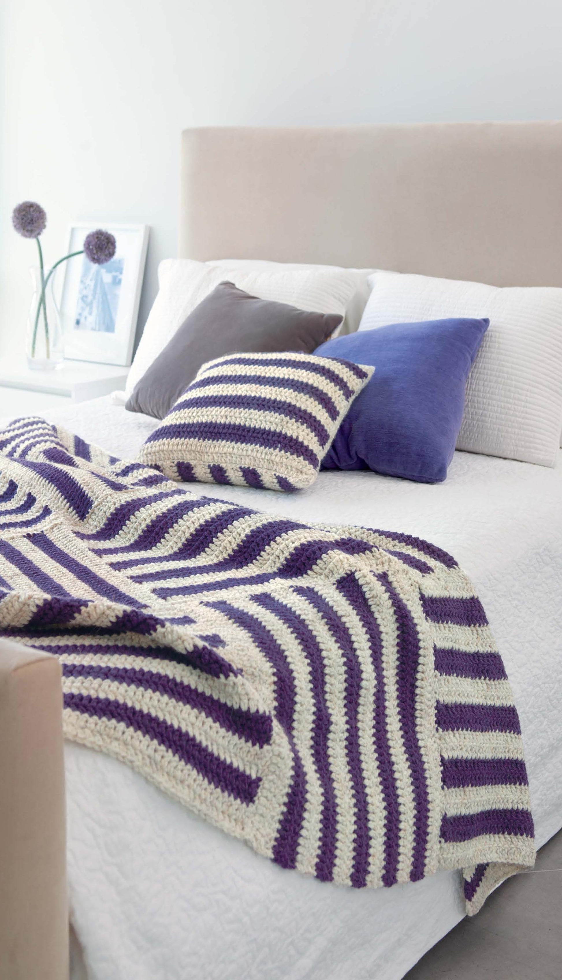 A neatly made bed features a cream and purple striped blanket and matching pillow from "Crochet For The Home" by Leisure Arts, plus solid purple and gray pillows. A white nightstand with framed photos and purple flowers completes the look.
