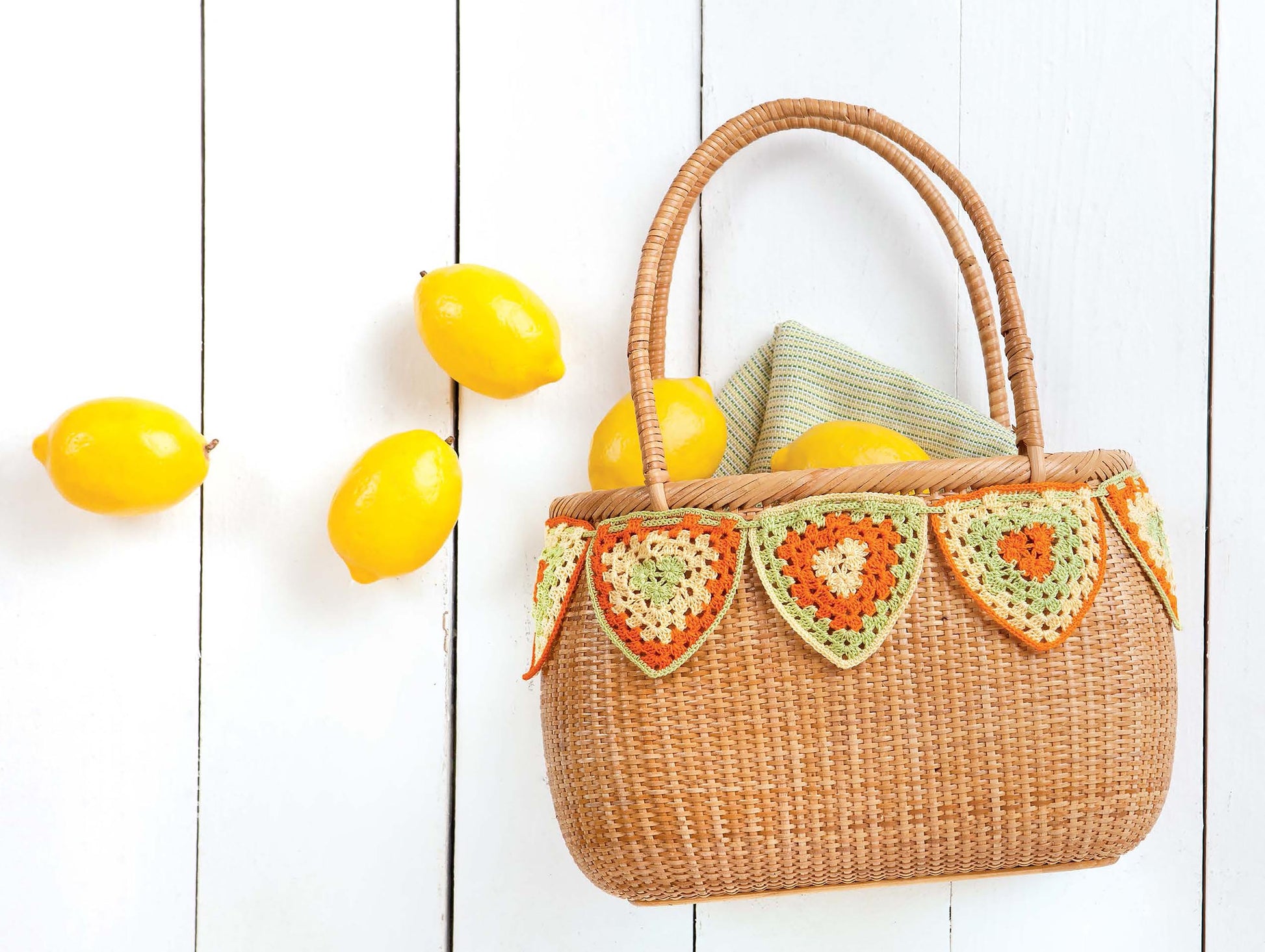 A woven basket with a green cloth and lemons inside is adorned with the "Crochet For The Home" garland by Leisure Arts, resting on a white wooden surface with several lemons scattered nearby.