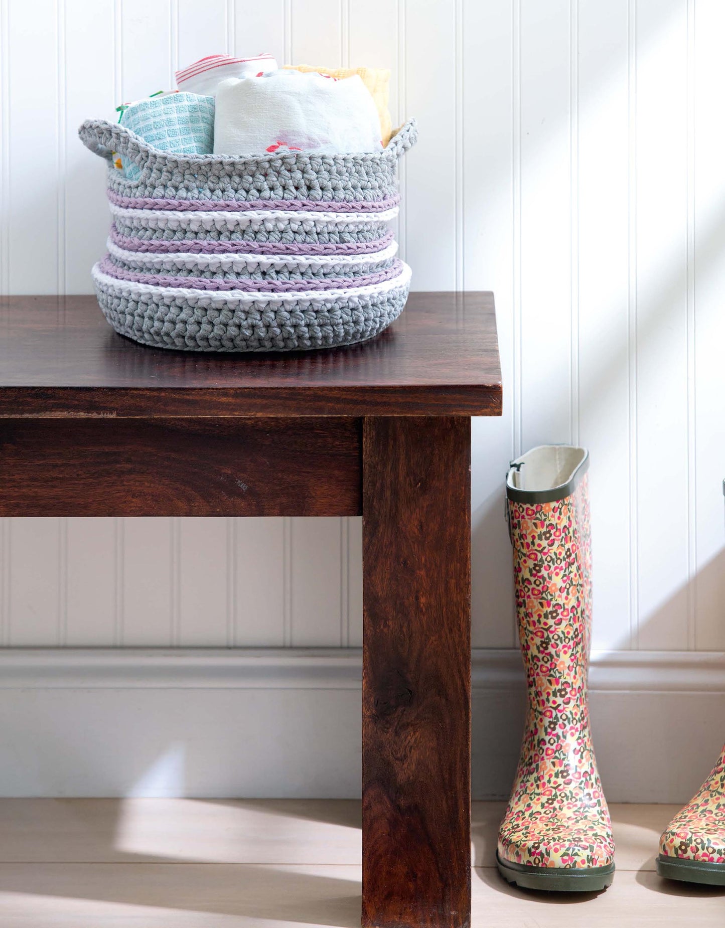 A wooden bench sits beside floral rain boots and a gray-lavender woven basket holding folded fabrics and crochet projects. Set against a white paneled wall, "Crochet For The Home" by Leisure Arts inspires creative home decor ideas.