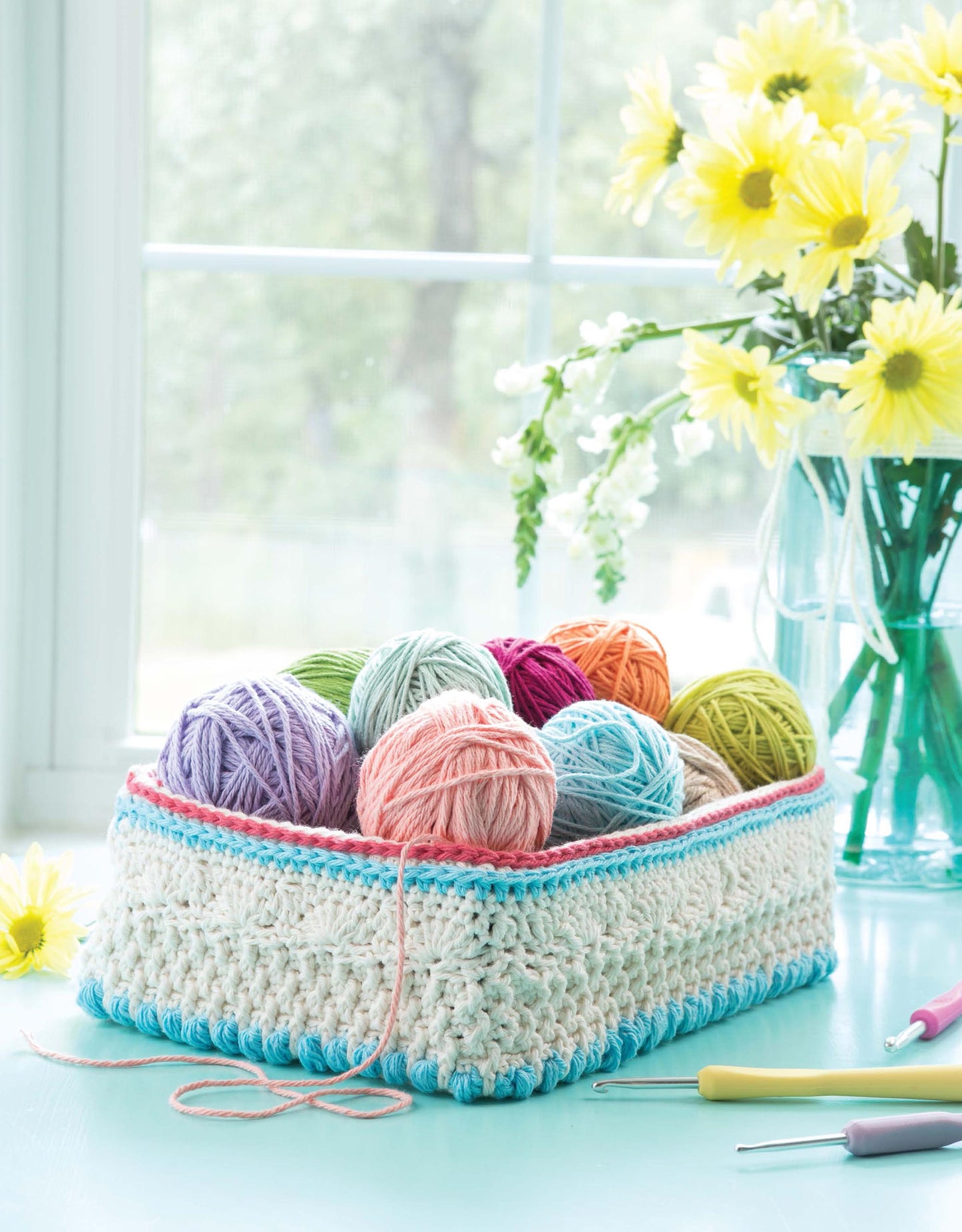 “Crochet For The Home” by Leisure Arts features a bright crocheted basket filled with colorful yarn on a blue table, accented by yellow flowers and hooks—ideal for cheerful home decor crochet in natural light.